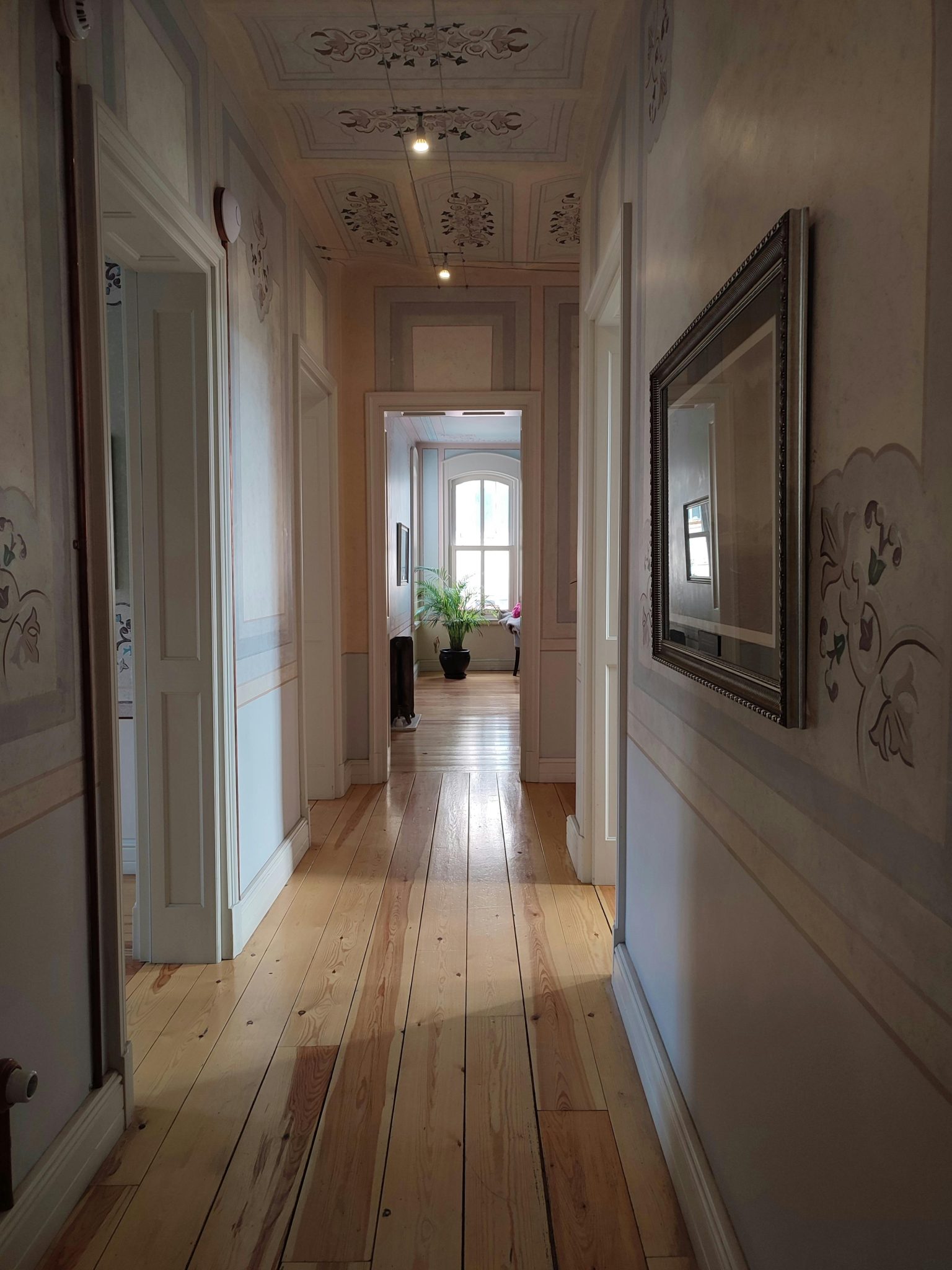 A beautifully lit corridor with wooden floors, ornate walls, and a plant at the end by the window.