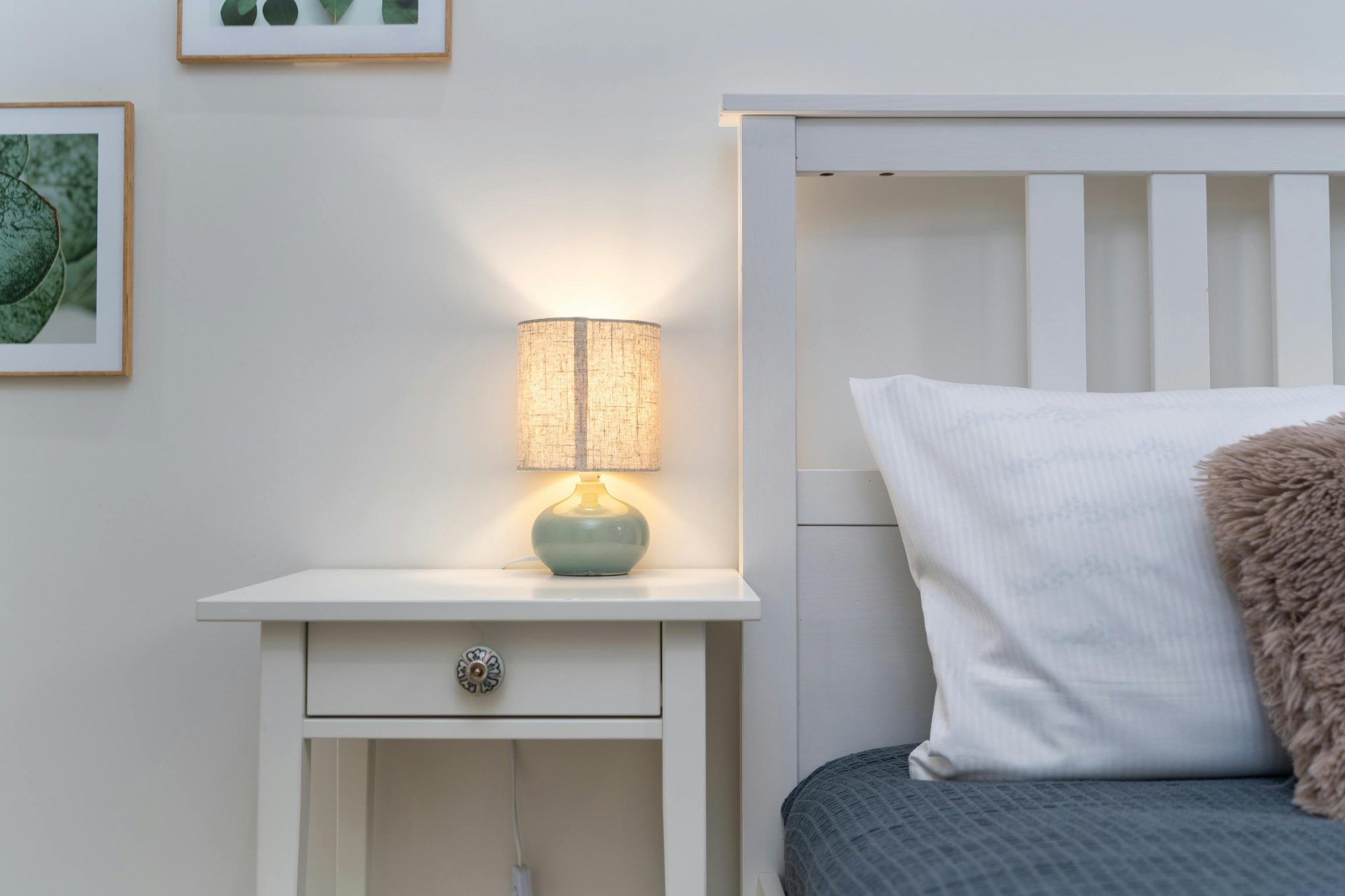A cozy bedroom setup featuring a lit lamp on a white nightstand beside a bed.