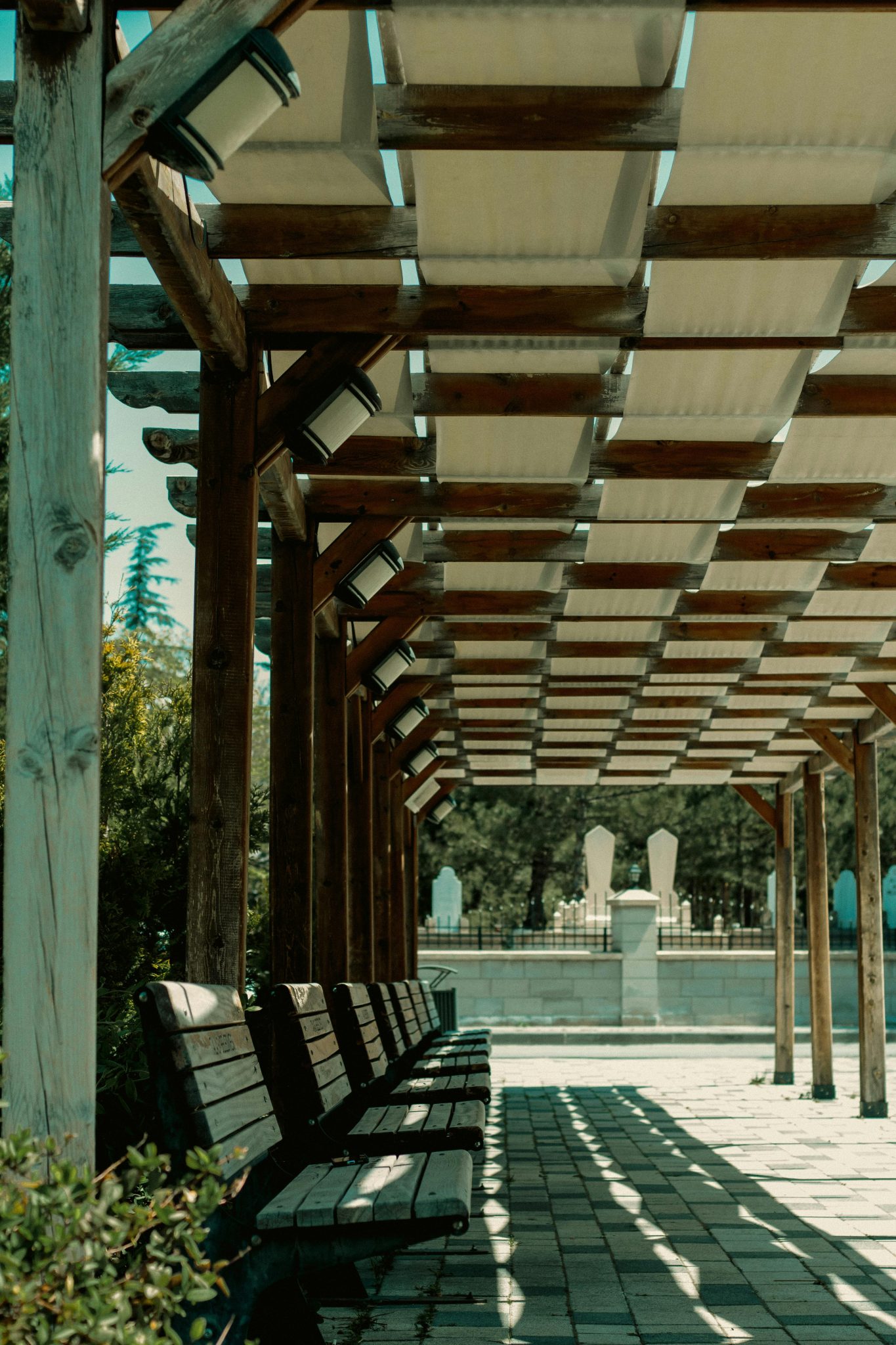 A serene outdoor seating area under a wooden pergola, casting detailed shadows in a sunny park.