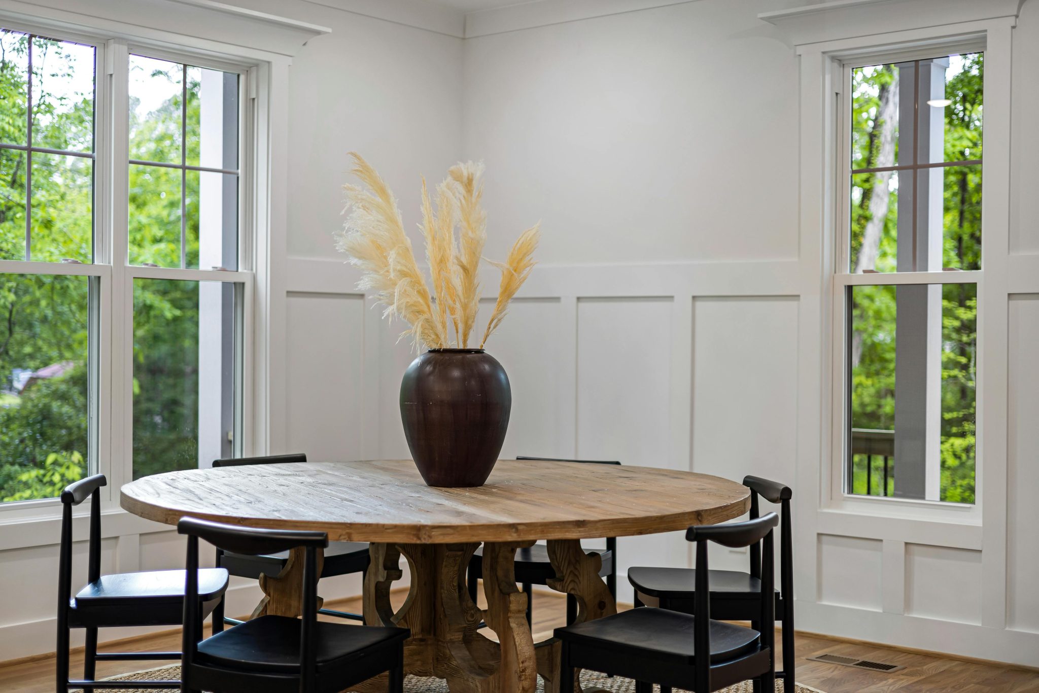 A stylishly decorated dining room featuring a round wooden table with a vase of pampas grass and black chairs.
