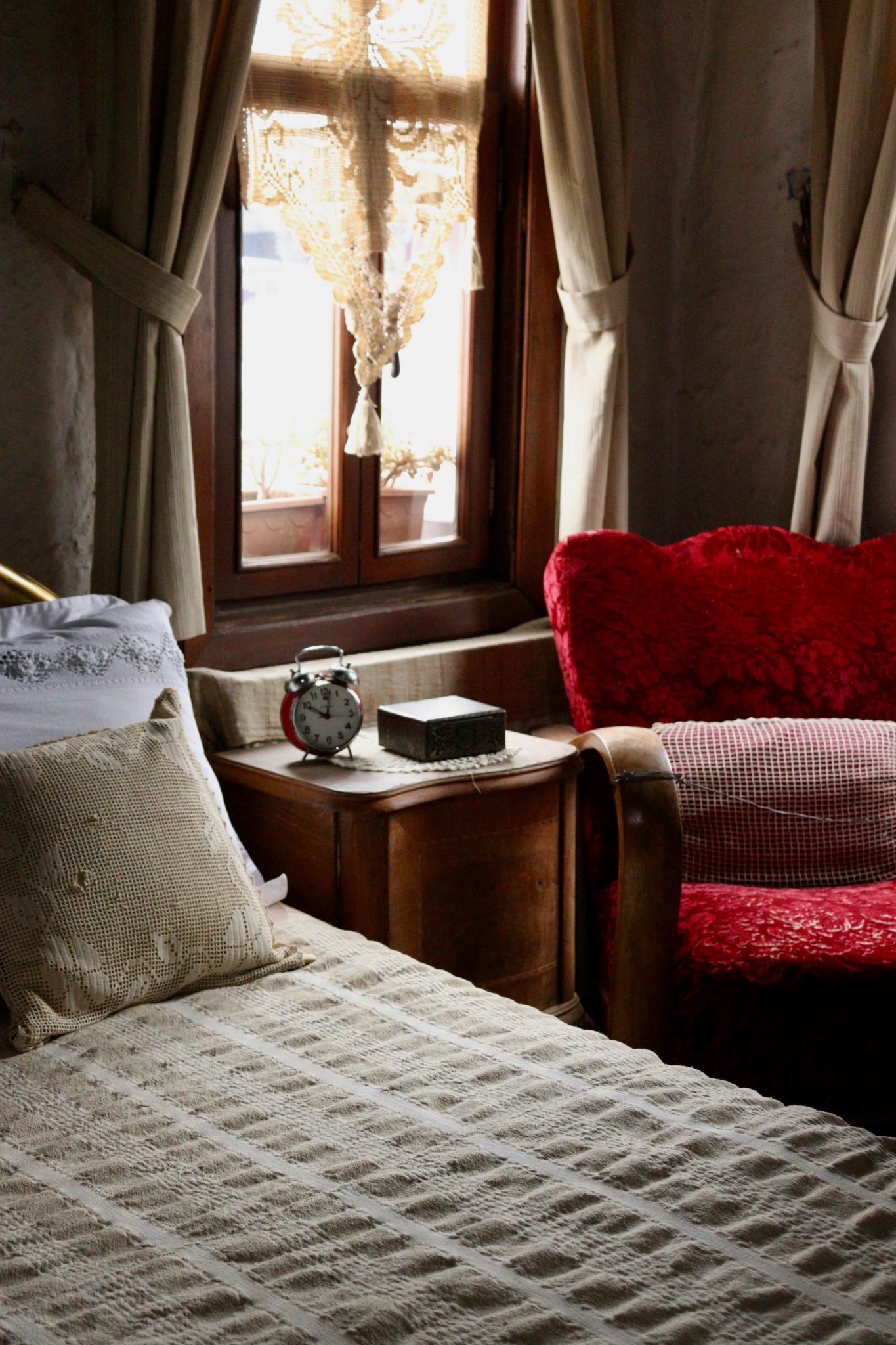 A vintage bedroom featuring a red armchair, lace curtains, and an antique alarm clock in warm lighting.