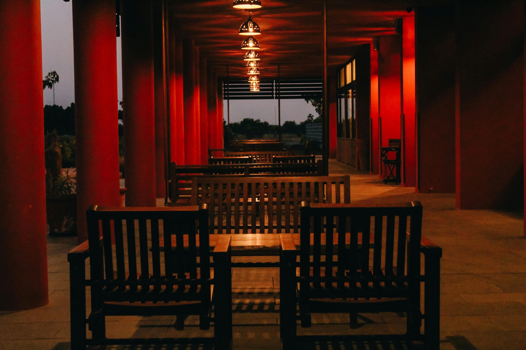 An outdoor wooden seating area with red columns, warmly lit in the evening.