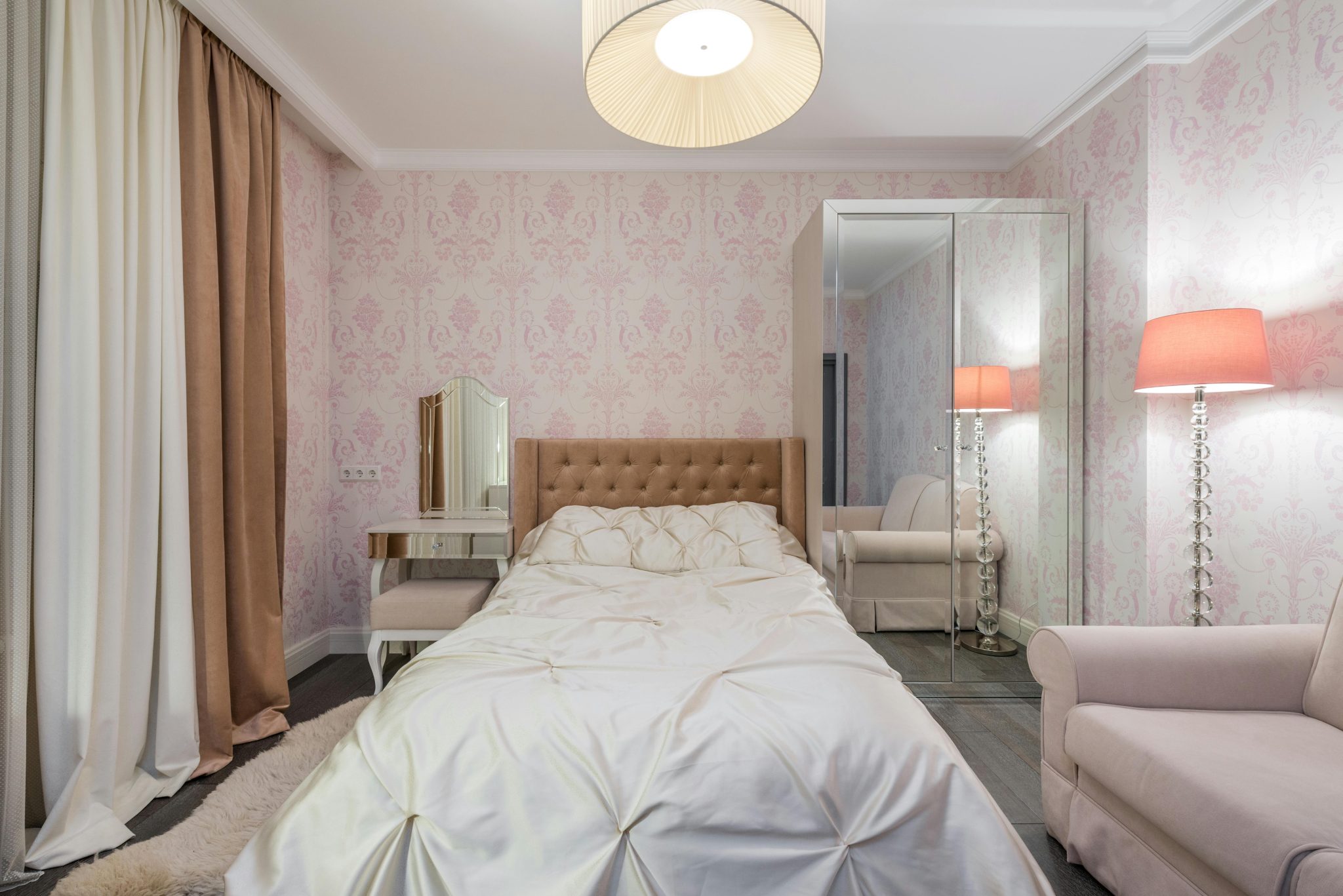Comfortable bed and soft couch in bedroom with pink walls and glowing lamps next to mirrored cupboard and curtains