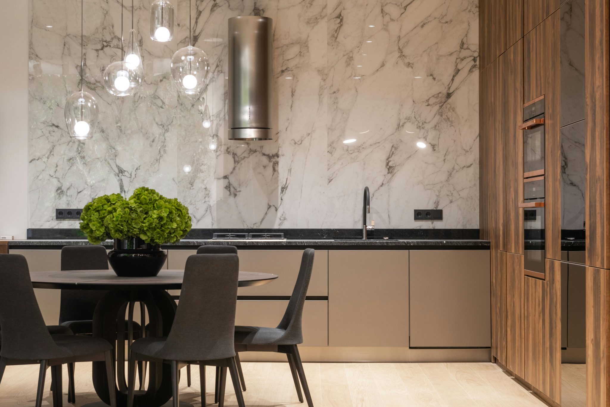 Comfortable chairs and round table with vase of fresh flowers in modern kitchen with wooden furniture and marble wall decorated with creative lamps