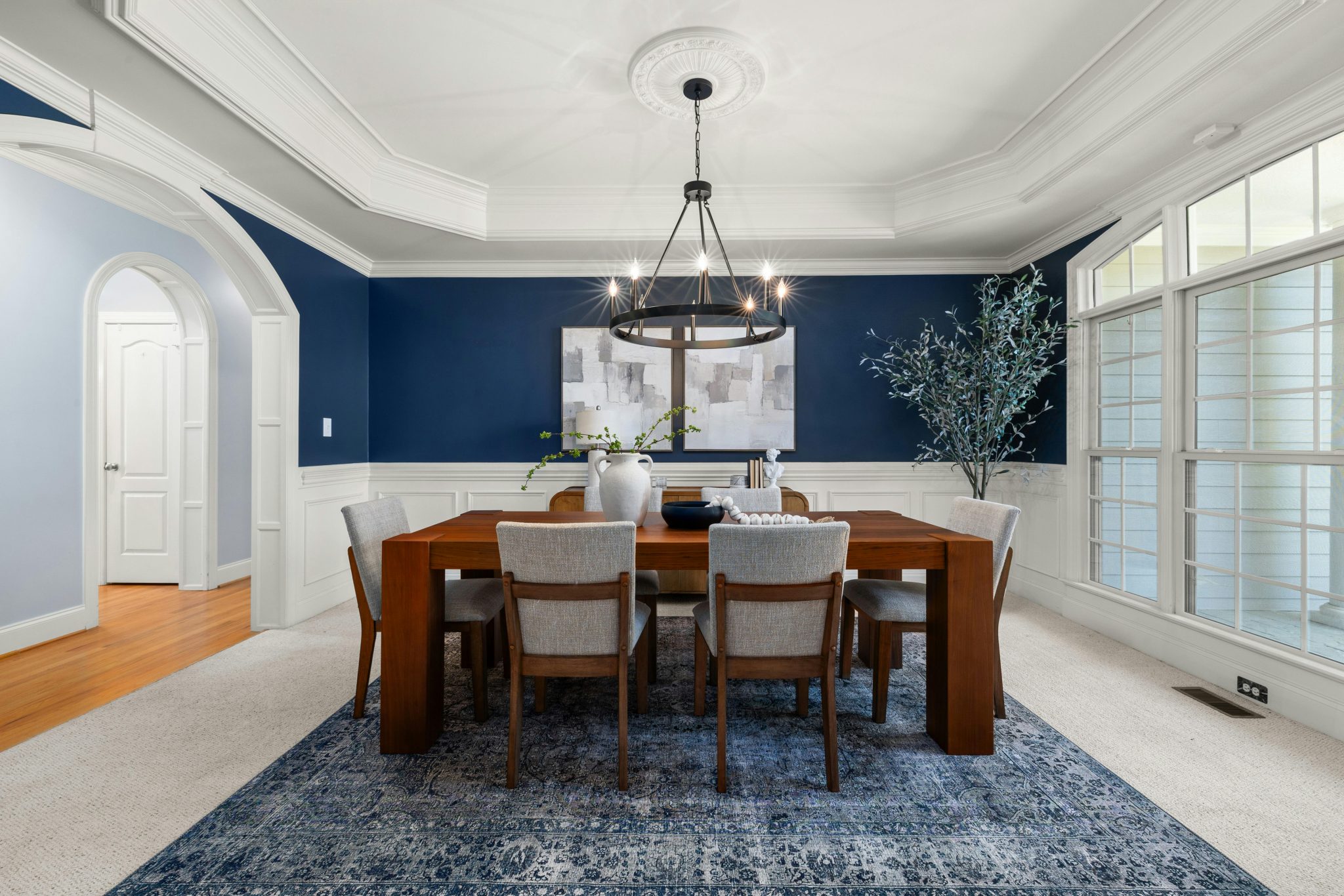 Contemporary dining room with wooden table, modern chairs, and elegant chandelier, featuring blue accent wall and large windows.