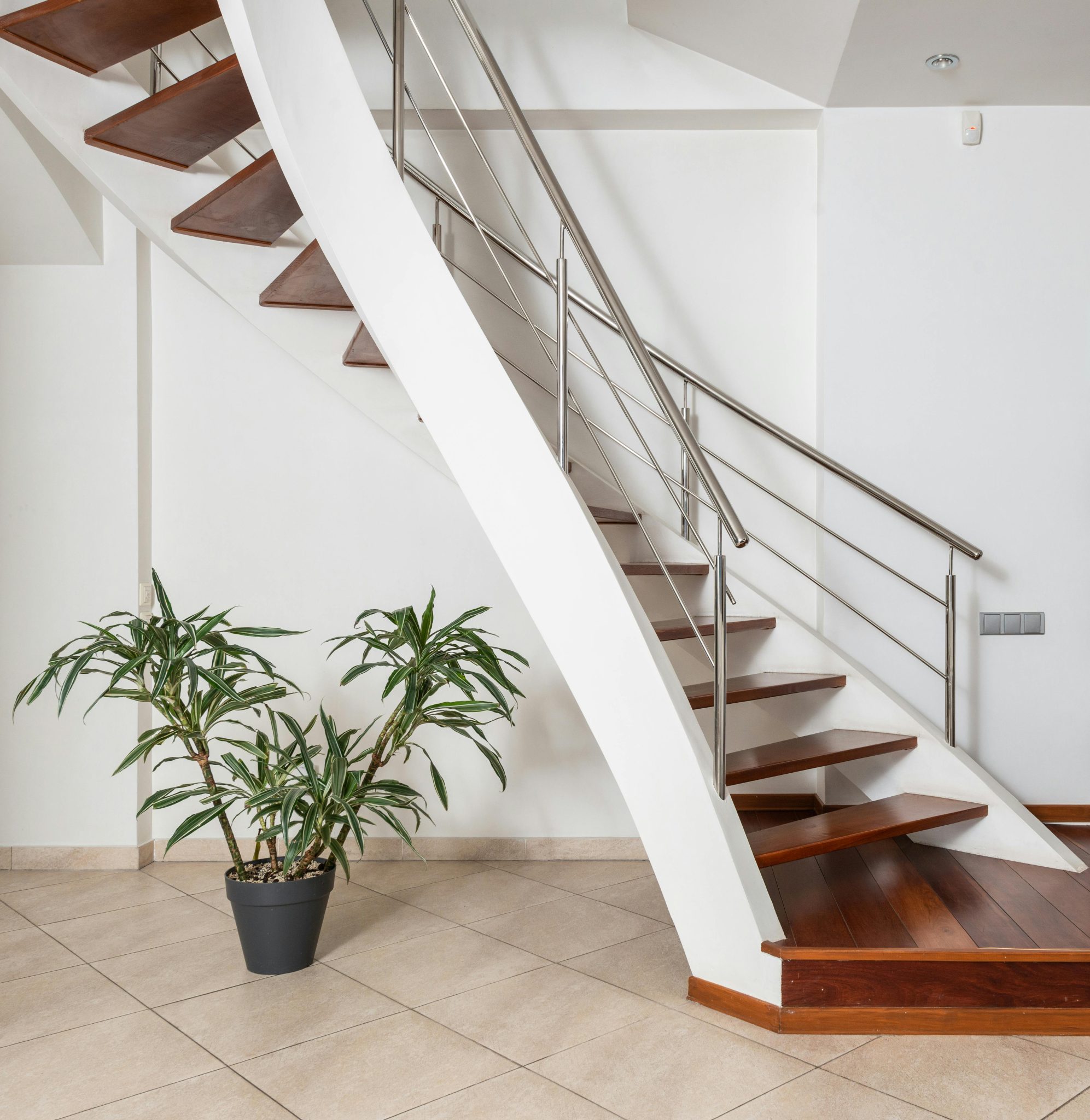 Contemporary wooden staircase with metal railings above ceramic floor with green plant in pot in light house