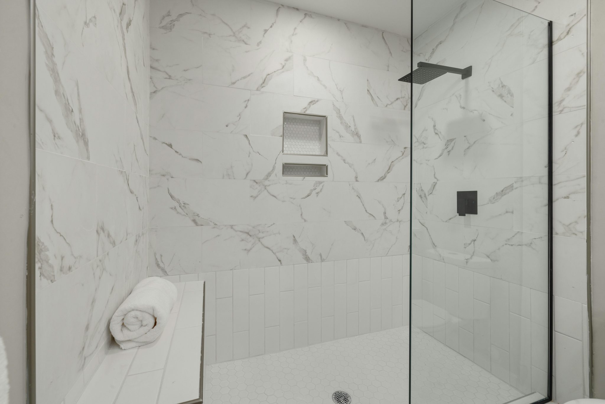 Elegant white marble shower with glass door, bench, and towel in modern bathroom.