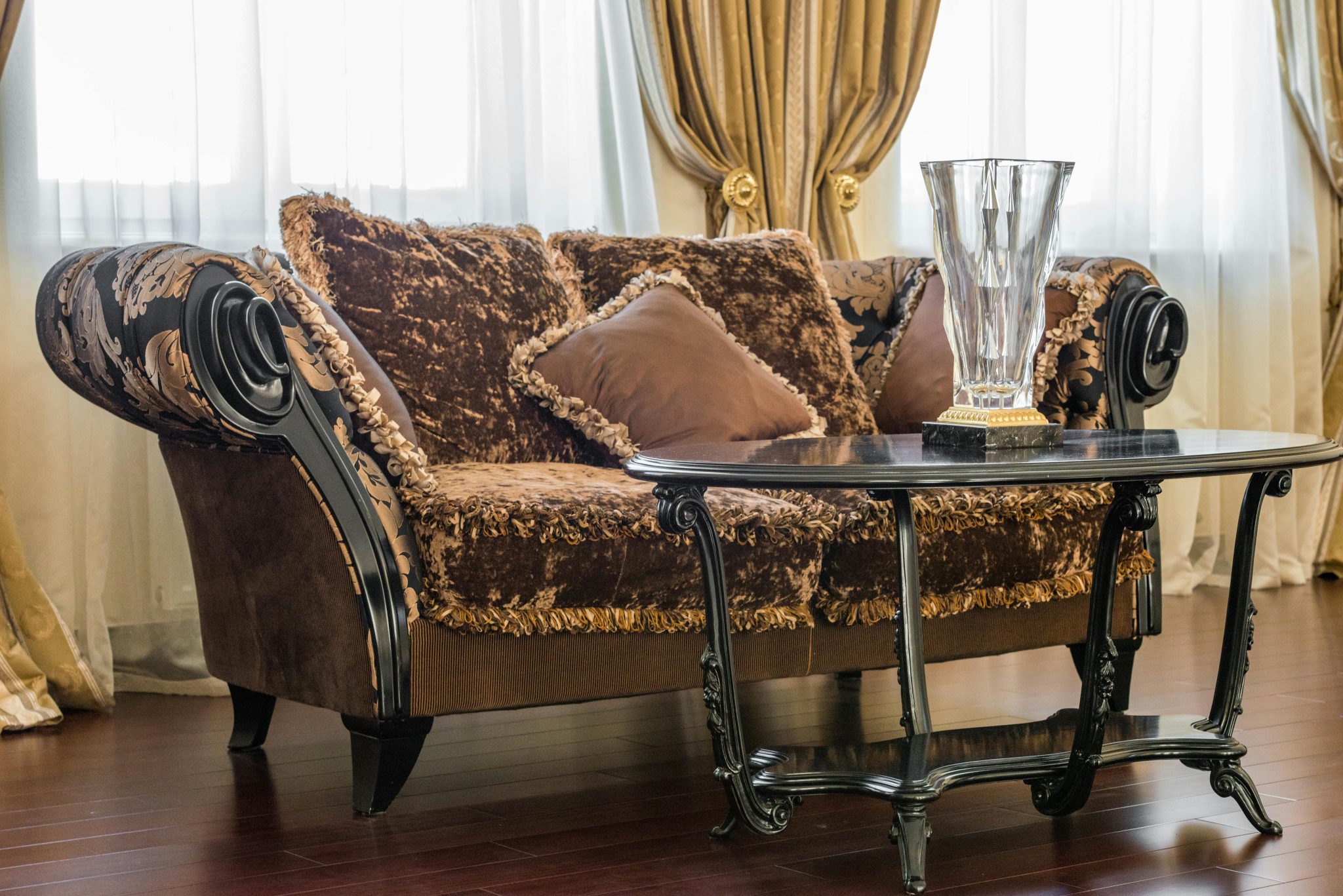 Interior of elegant living room with brown couch with pillows near table with glass vase next to curtains on windows in daylight