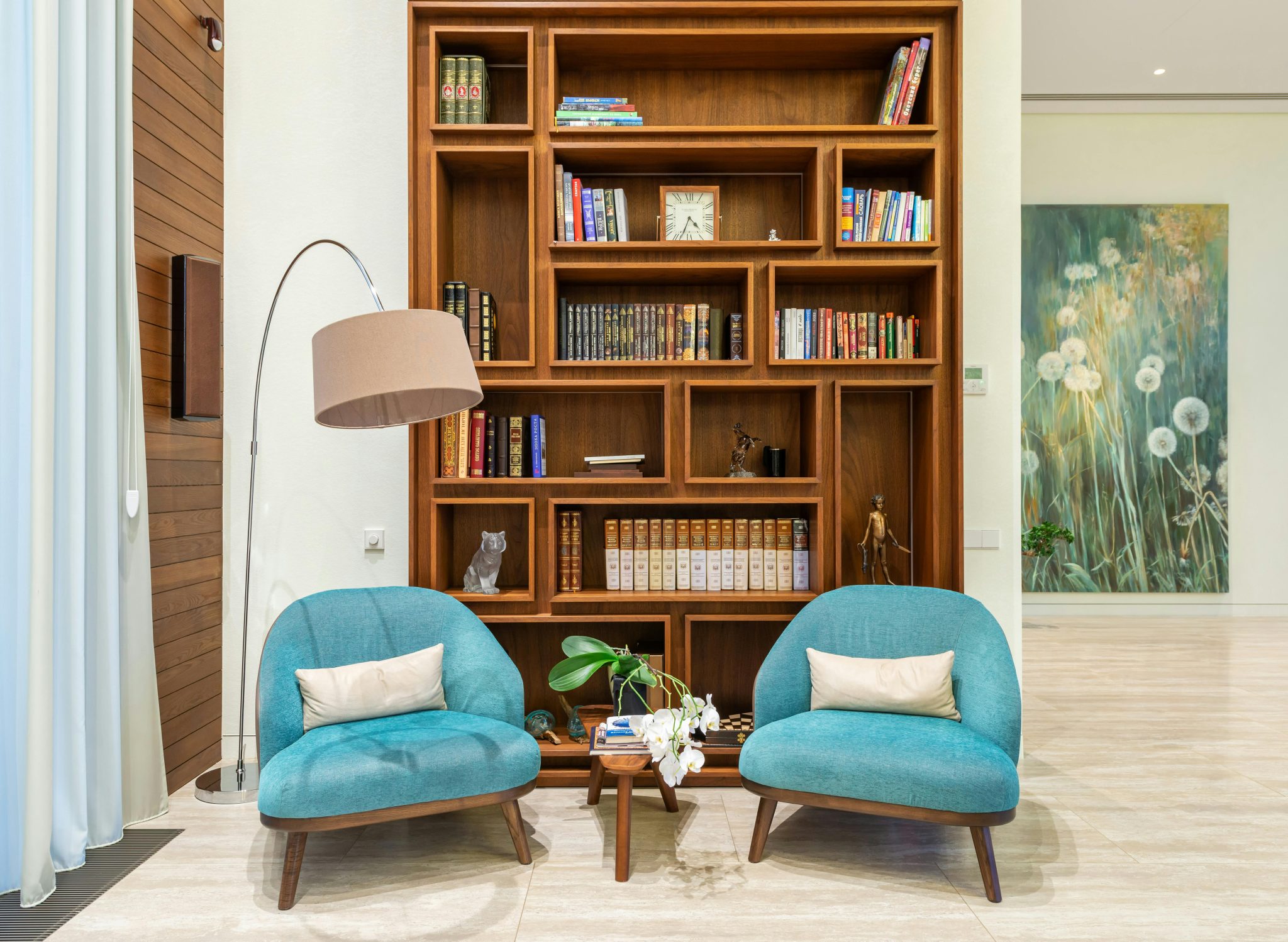 Interior of living room with cozy soft blue armchairs near lamp and books placed on shelves of wooden bookcase