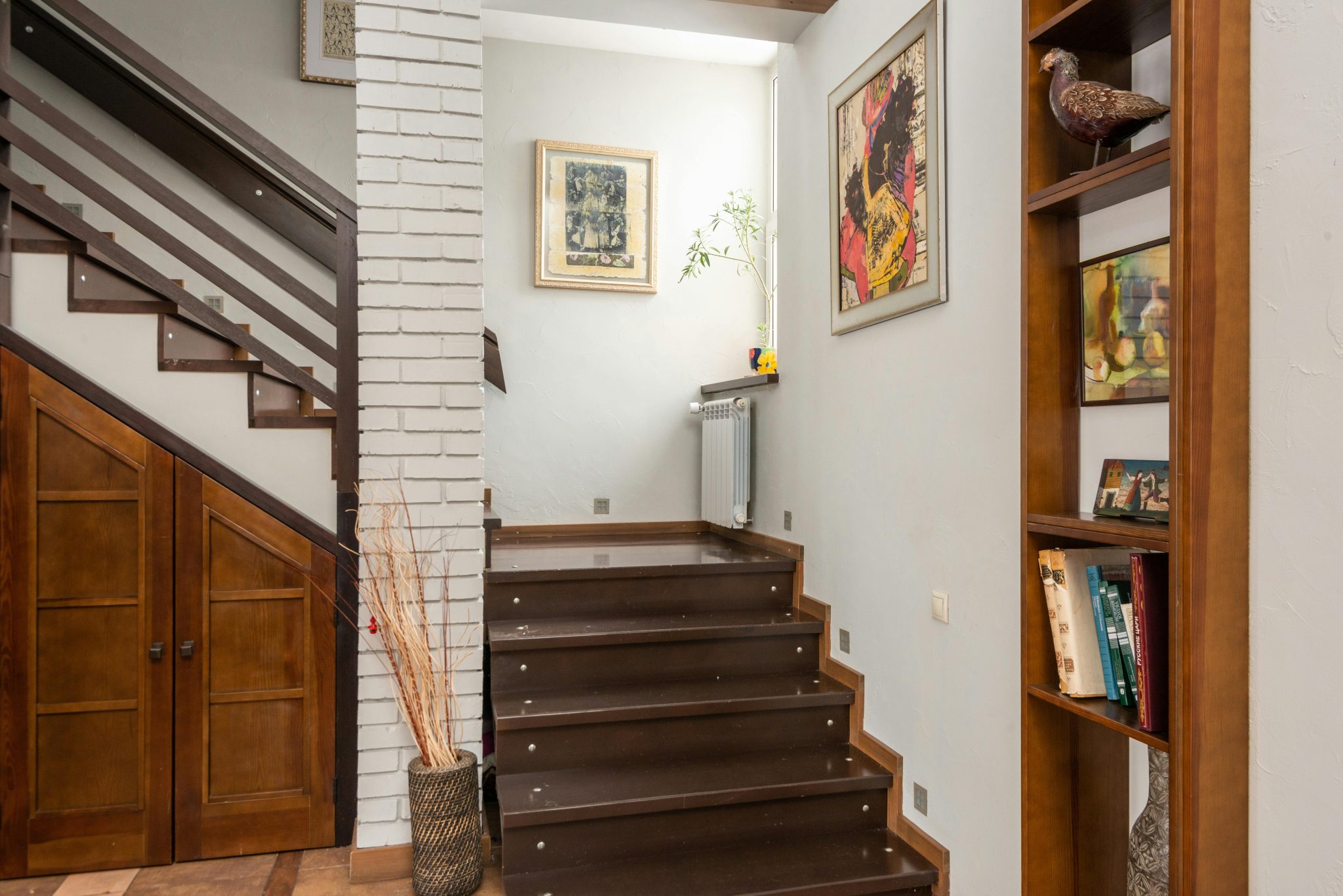 Interior of modern flat with stairway placed near brick column and wooden shelf with decorative elements at wall with paintings