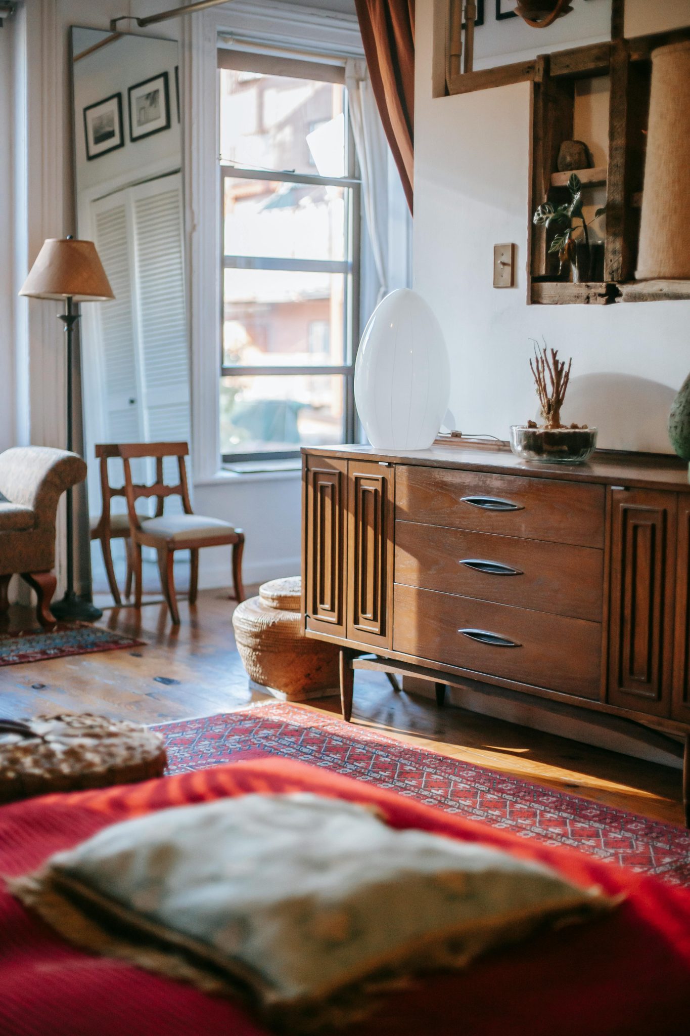Light spacious bedroom interior furnished with vintage chairs and wooden retro dresser with drawers opposite bed covered with red blanket on sunny day