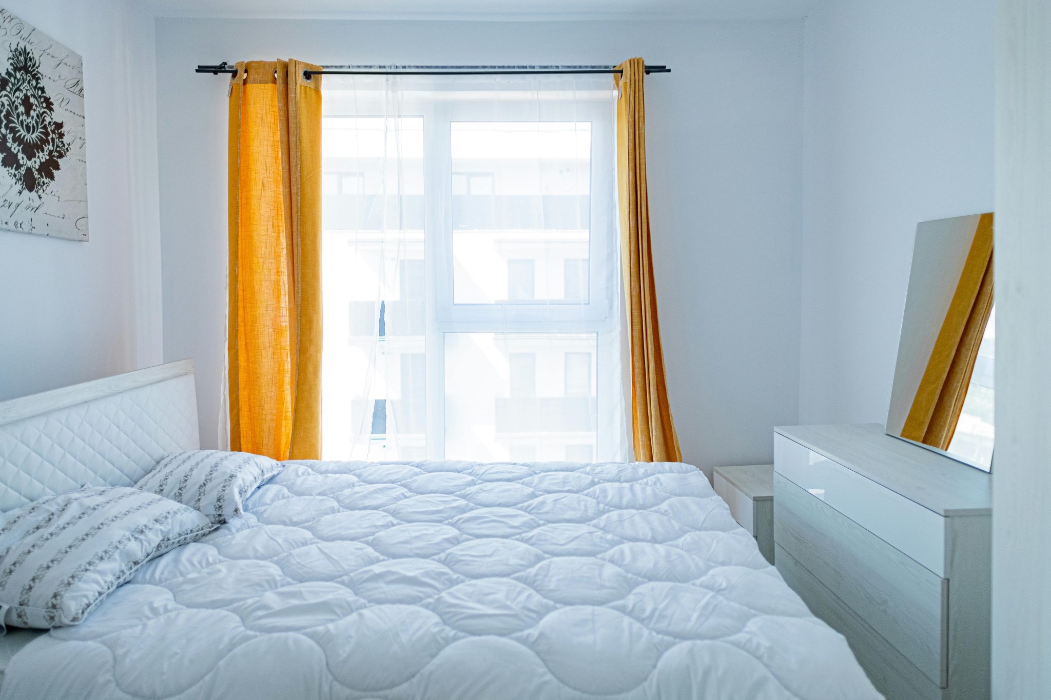 Minimalist bedroom featuring a white bed, mirrored dresser, and yellow curtains for a fresh look.