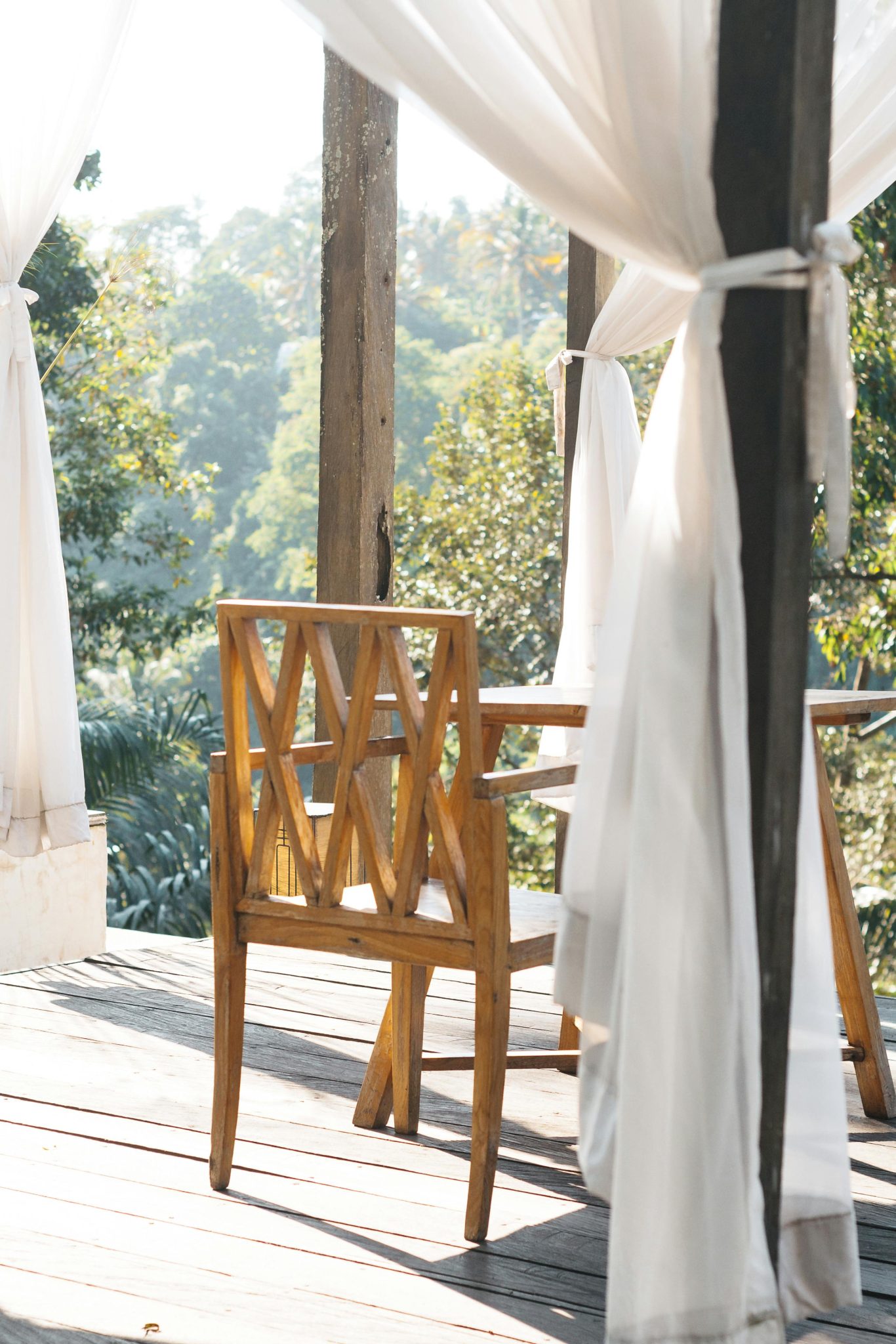 Relaxing outdoor patio with wooden chair and flowing curtains in a natural setting.