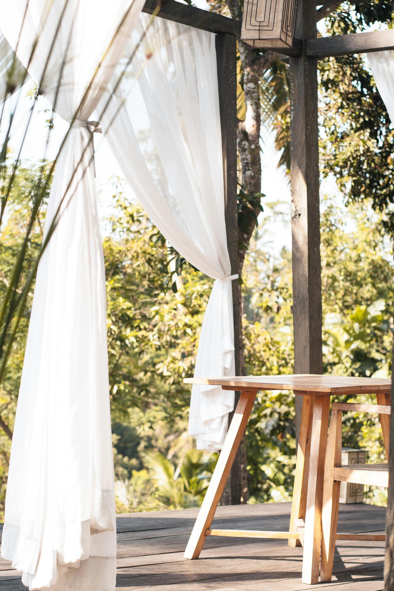 Serene outdoor patio with white curtains and wooden table, nestled among green trees. Ideal for a relaxing escape.