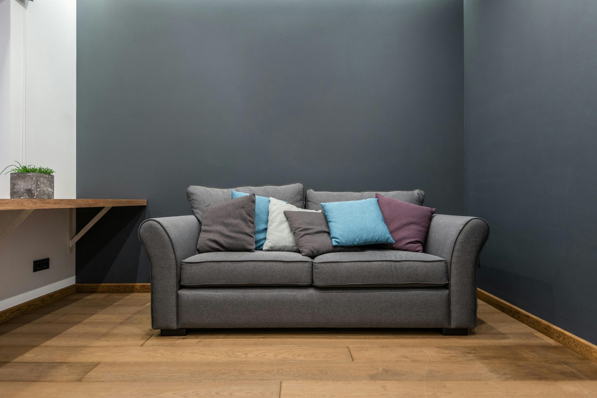 Soft gray sofa with abundance of colorful cushions placed near wall in light room with potted plant on wooden shelf
