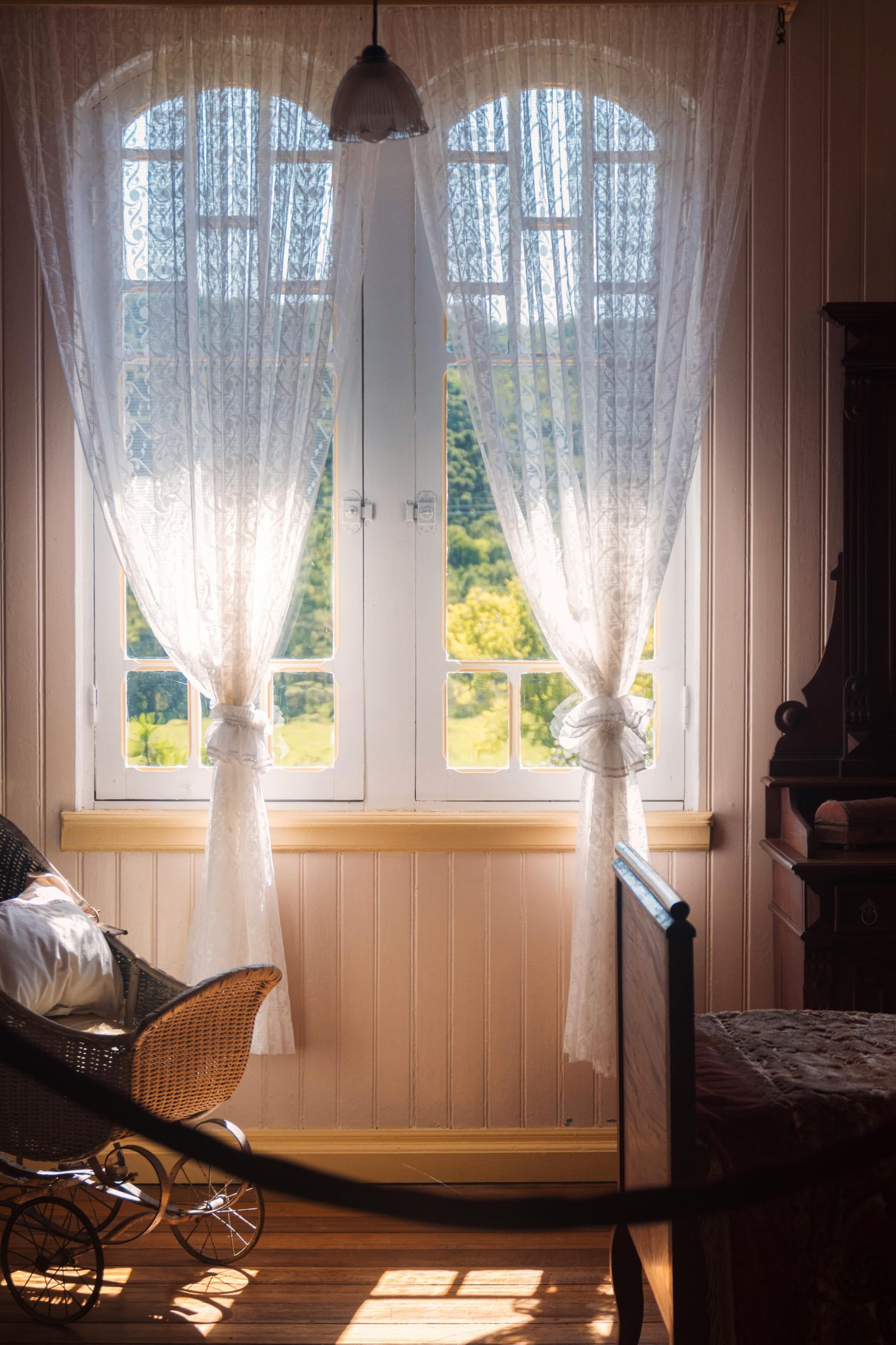Sunlight streams through lace curtains in a vintage bedroom with a view of lush greenery.