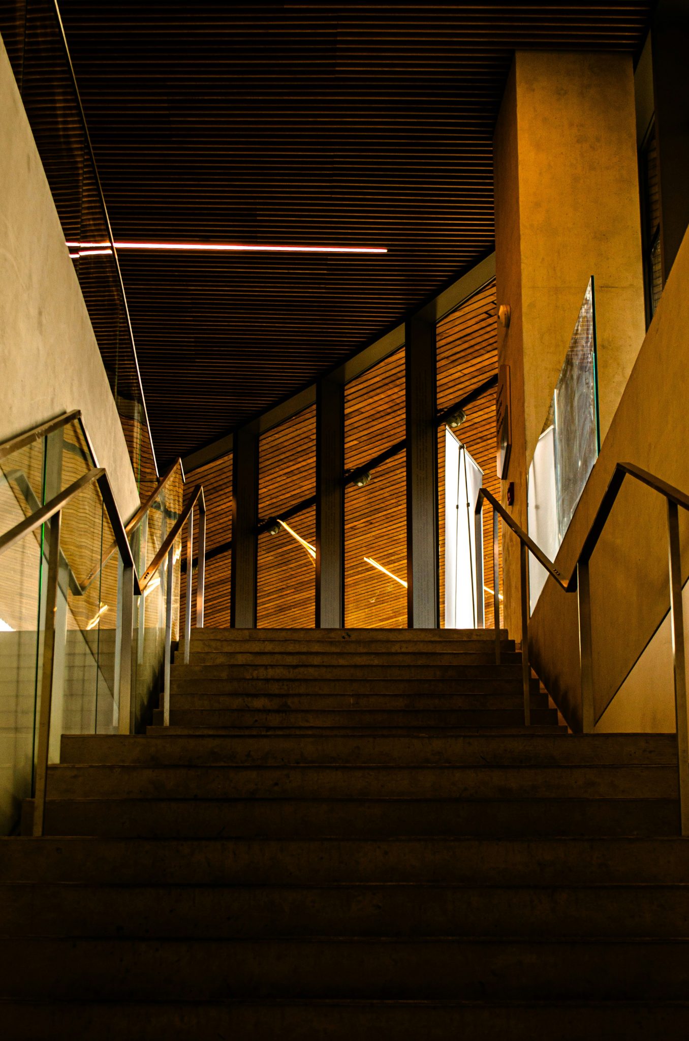 The image depicts a modern, architecturally striking staircase leading upward within an indoor space. The composition emphasizes clean lines, geometric shapes, and contrasting textures. The warm woode