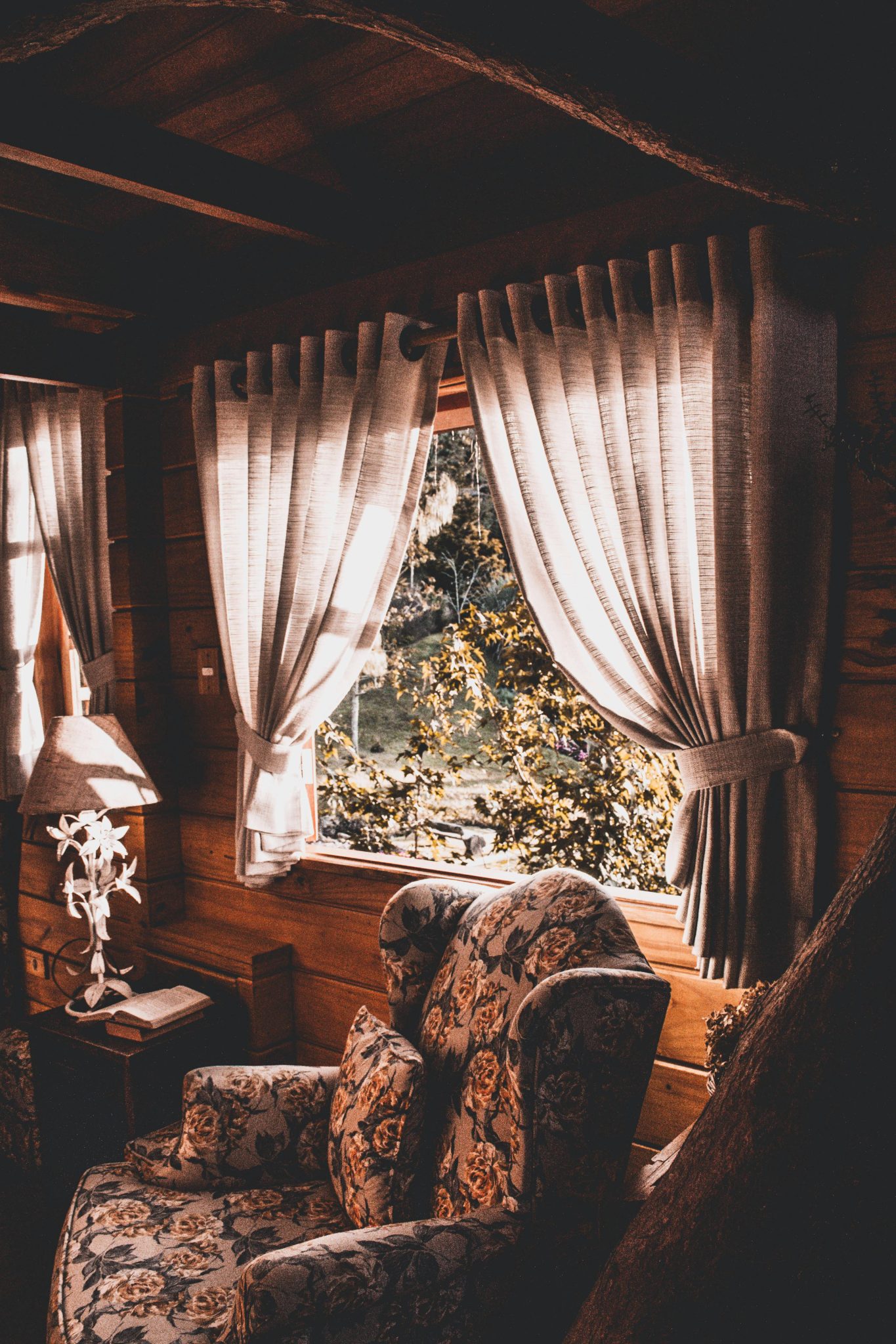 Warm and inviting rustic living room with floral armchair, wooden walls, and sunlight filtering through curtains.