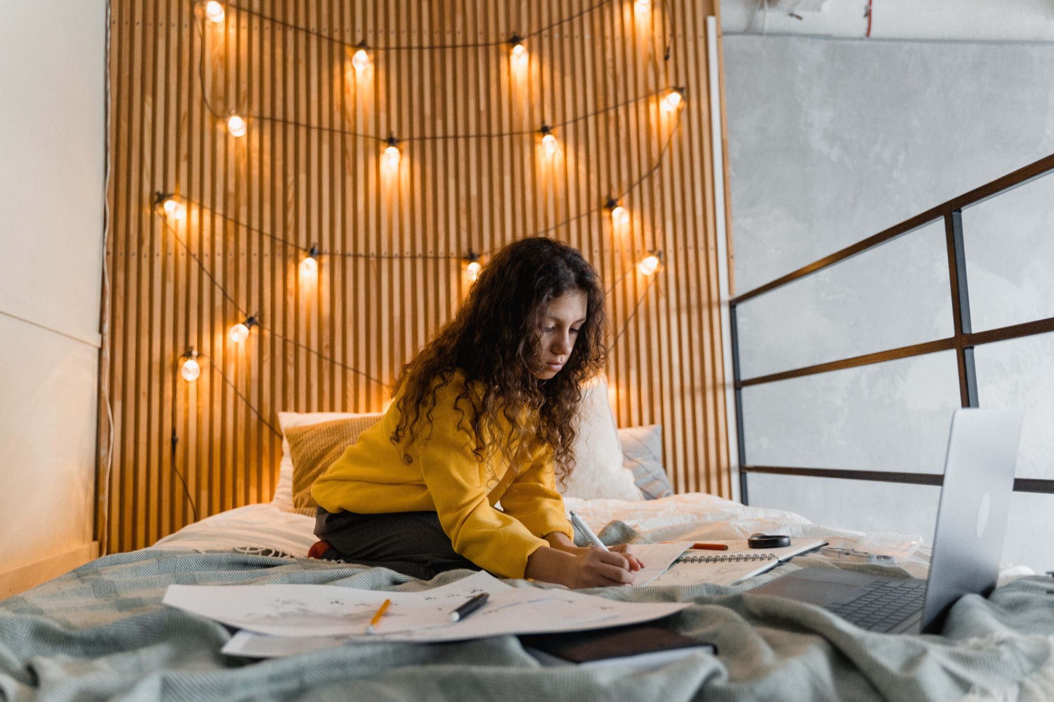 Woman writing on bed with laptop in a cozy bedroom ambiance with string lights.