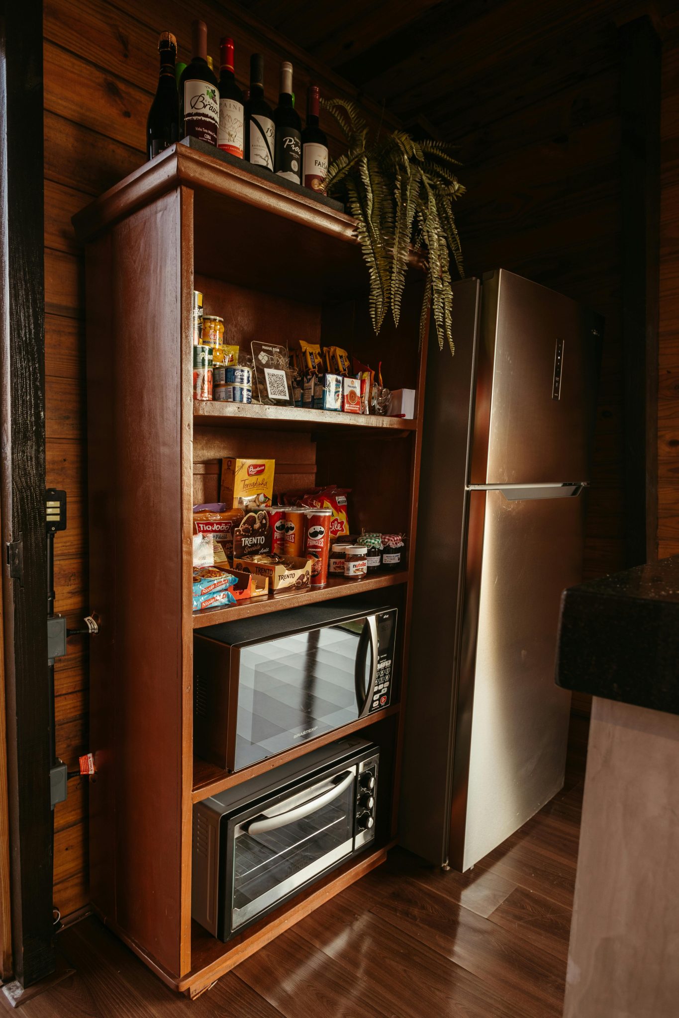 Wooden shelf with snacks and wine, next to modern fridge, in a rustic kitchen setting.