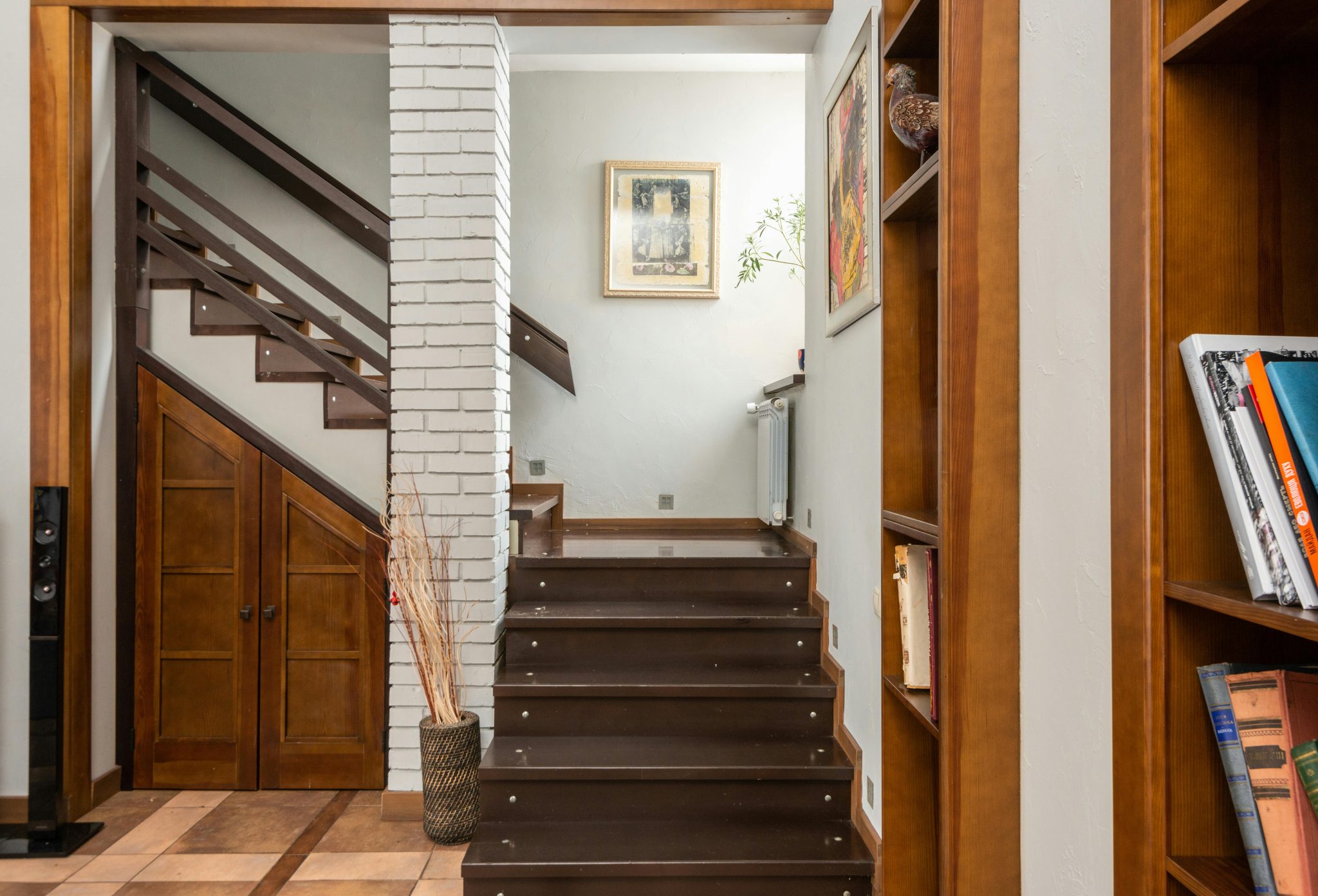 Wooden stairway near brick column placed at shelves with books in modern flat with pictures on wall and dry plants in vase