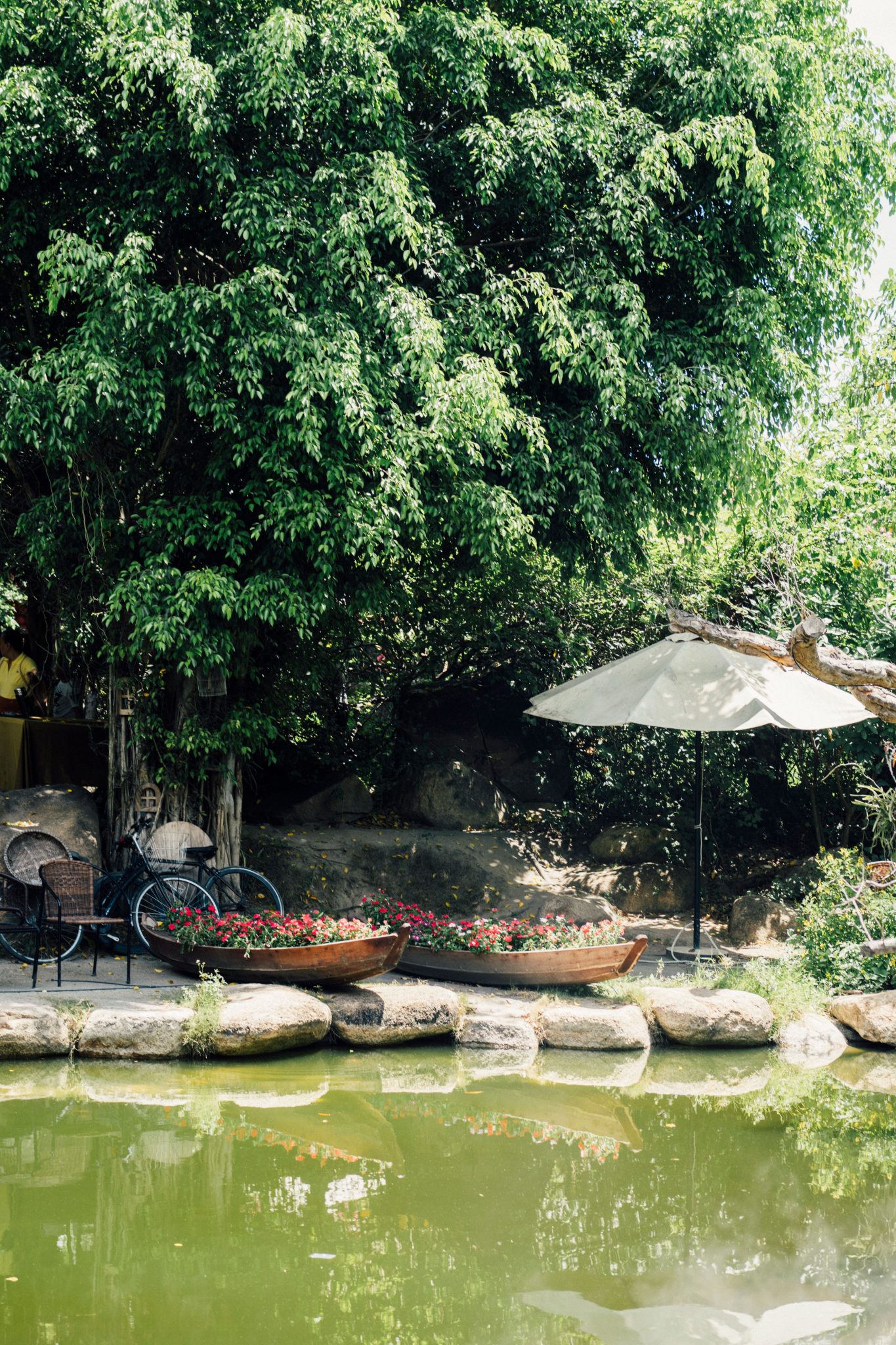 A serene garden scene featuring a pond, trees, chairs, and an umbrella, ideal for outdoor relaxation.