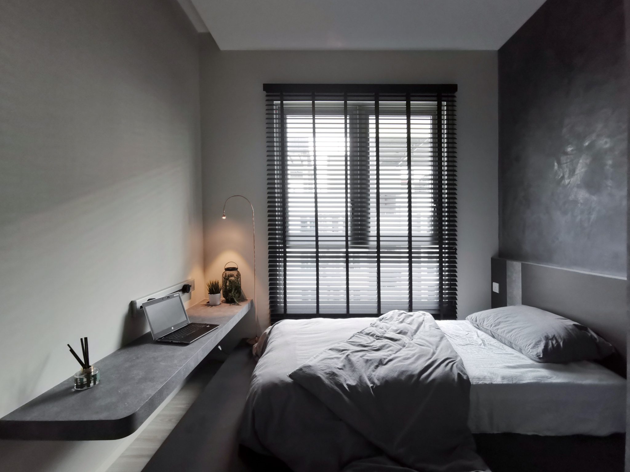 A stylish and minimalist bedroom with natural light streaming through the blinds, featuring a cozy bed and workspace.