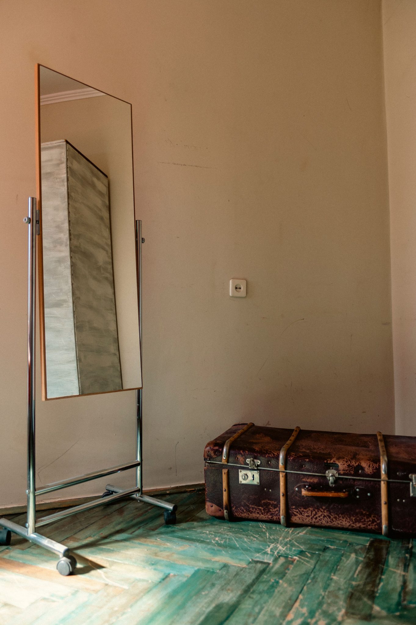 A vintage suitcase rests against a wall with a tall mirror reflecting light in a rustic room.