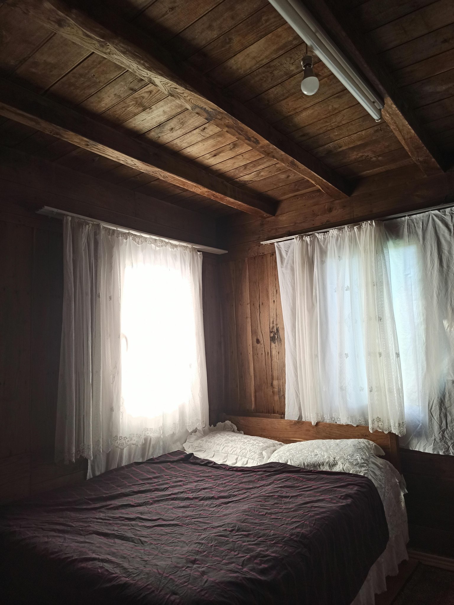 A warmly lit rustic bedroom featuring wooden walls, ceiling, and soft white curtains.