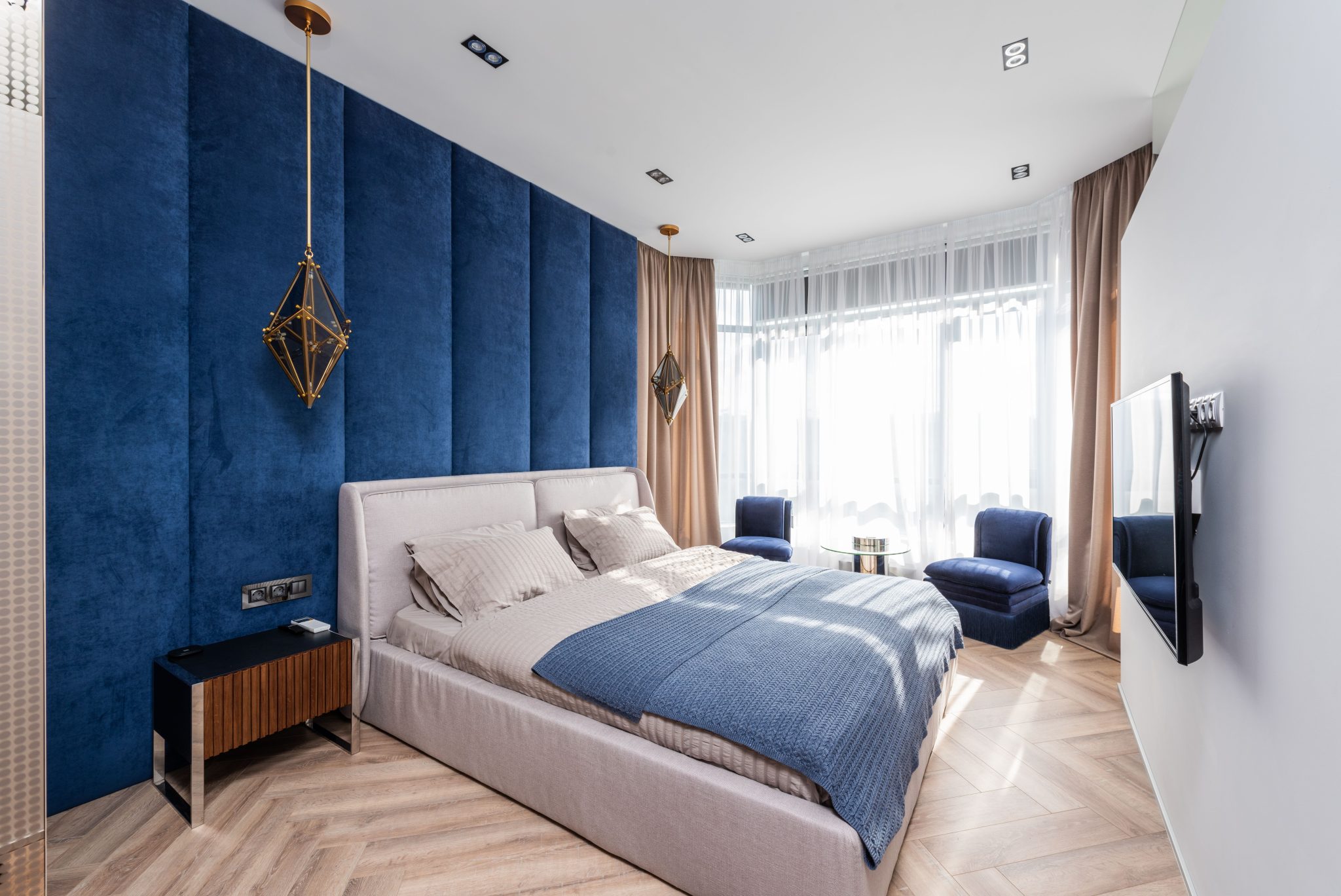Comfortable bed with cushions and blanket placed near blue wall against TV in spacious bedroom with armchairs and table near window