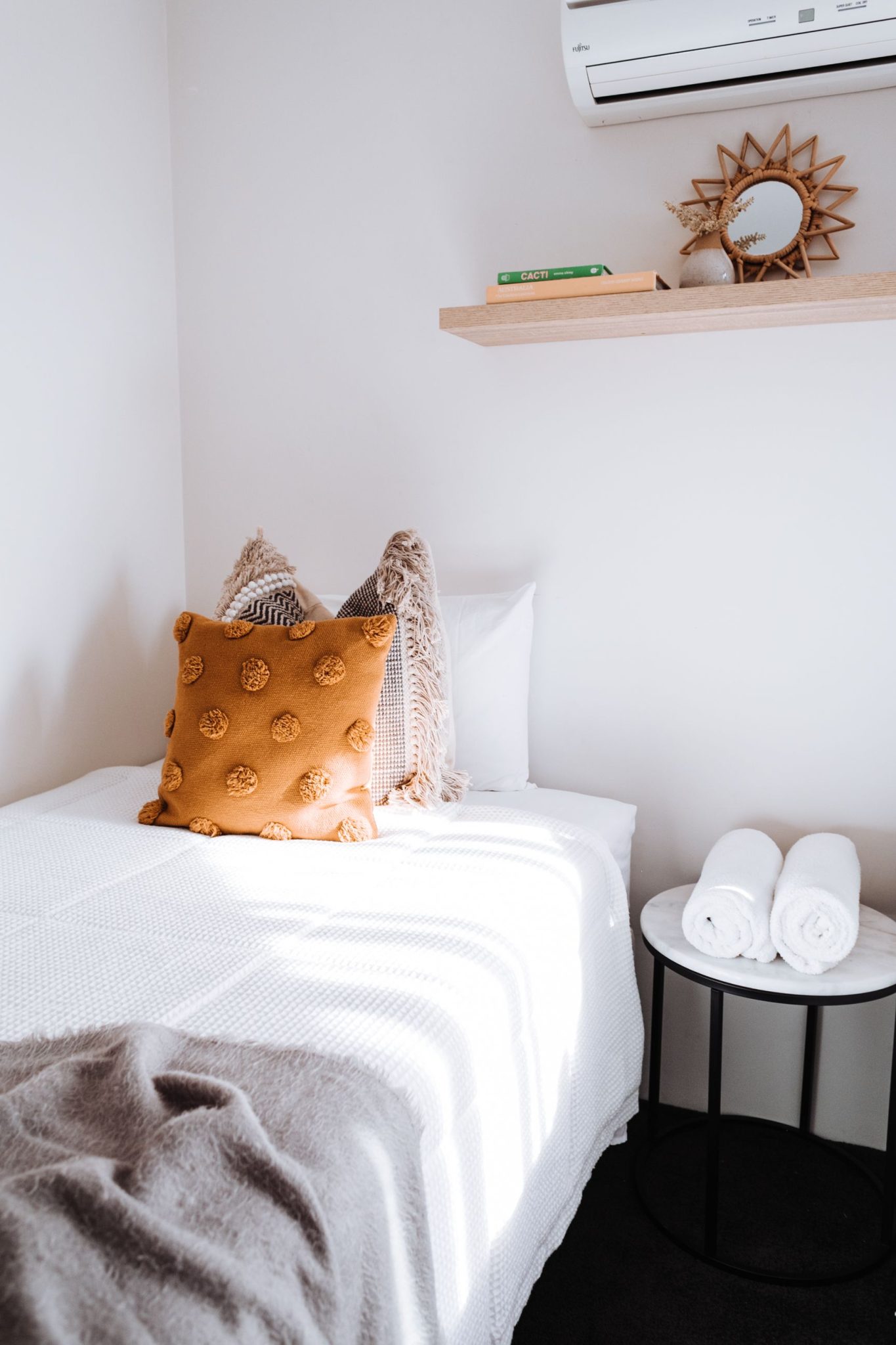 Comfortable bed with soft beige cushions and cotton plaid in modern bedroom with air conditioning book shelf and towels on bedside table