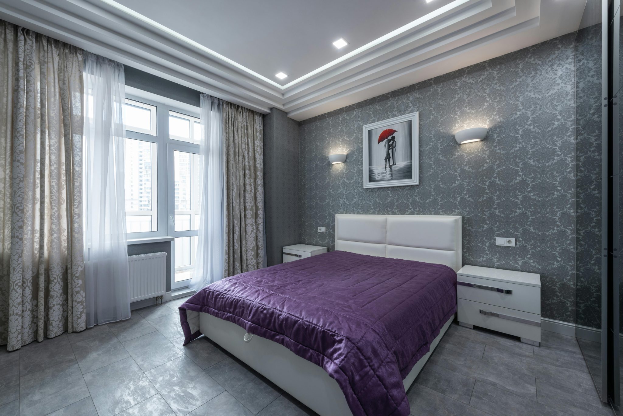Comfortable white bed with purple coverlet placed in stylish bedroom with gray walls in modern flat