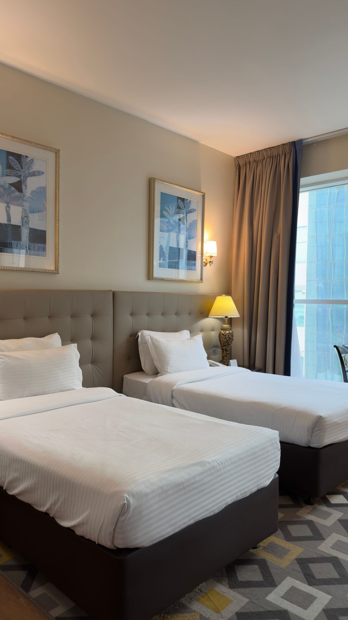Cozy and modern hotel room featuring twin beds, elegant decor, and a stunning city view through large windows.