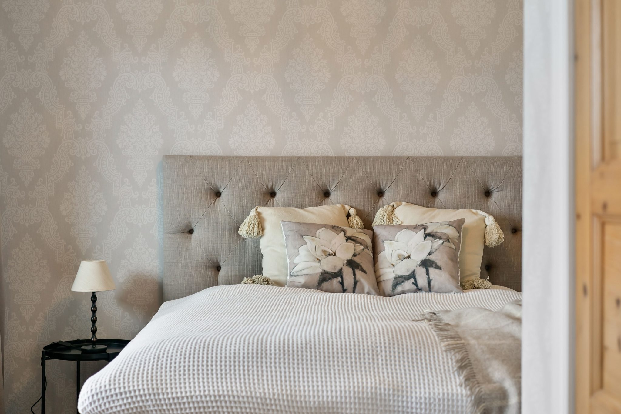 Elegant bedroom interior with floral pillows, beige wallpaper, and soft textiles creating a cozy ambiance.