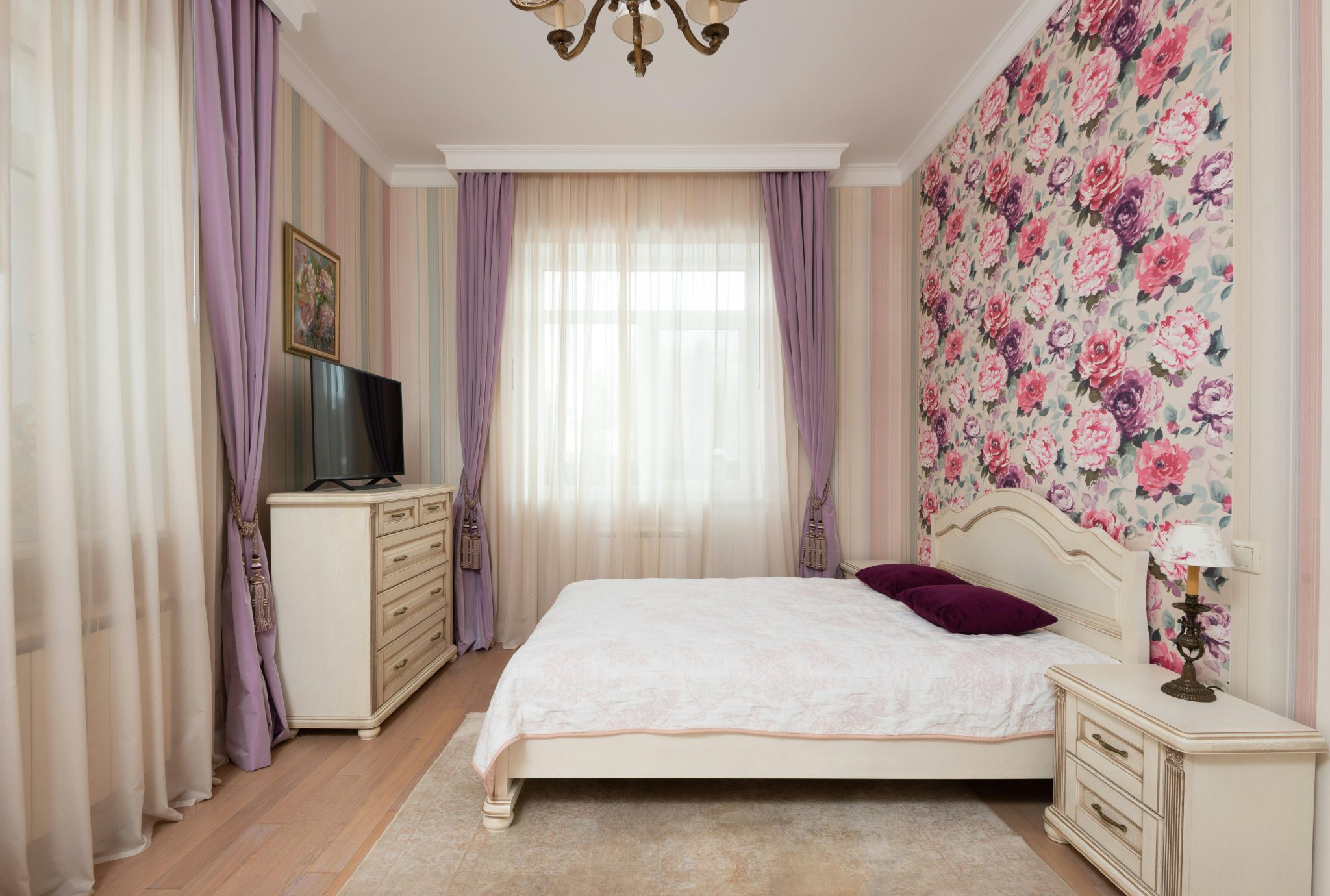 Interior of light bedroom with soft bed in front of chest of drawers with TV near windows with light curtains