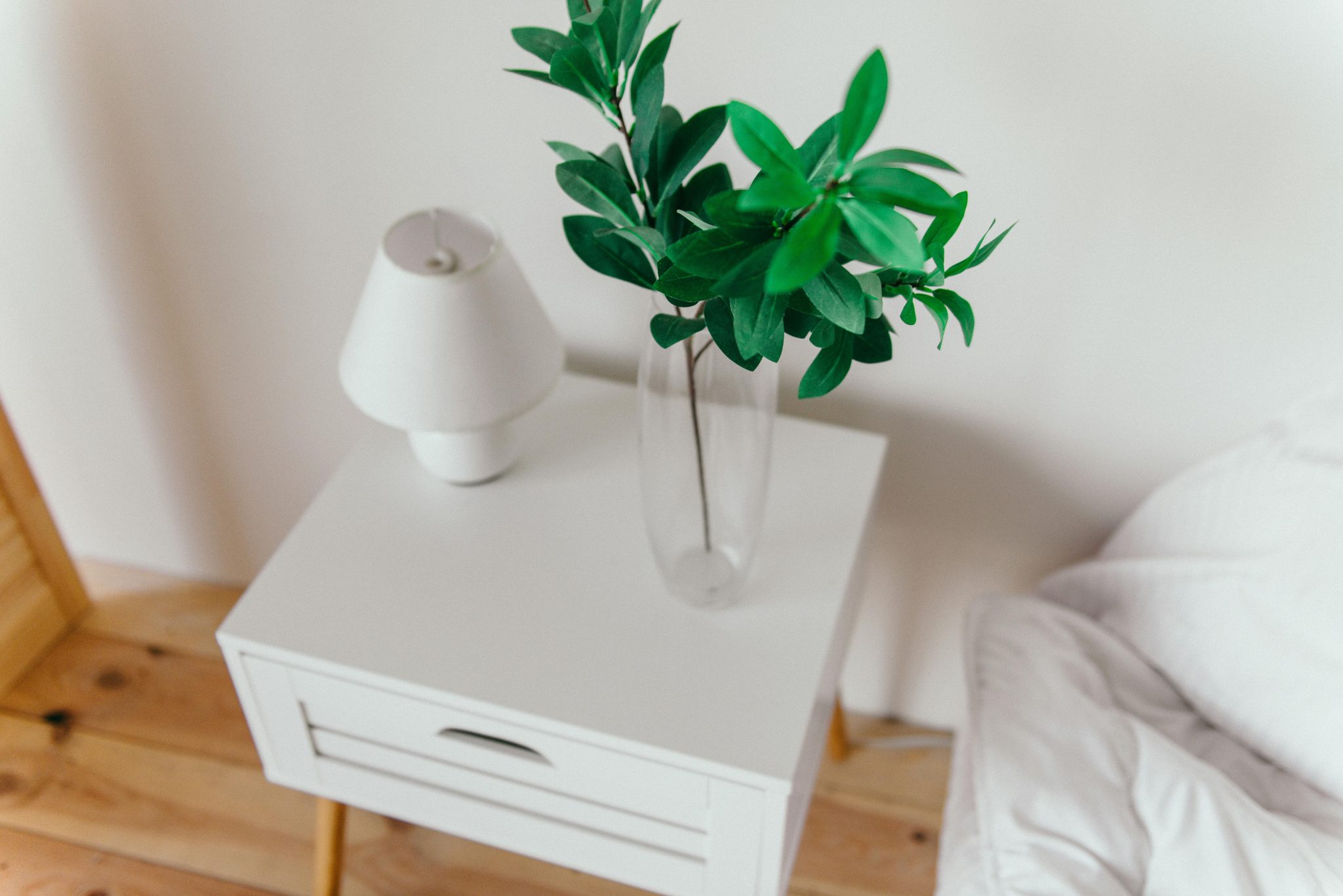 Modern bedroom interior featuring a stylish white side table, sleek lamp, and a vase with fresh green leaves.