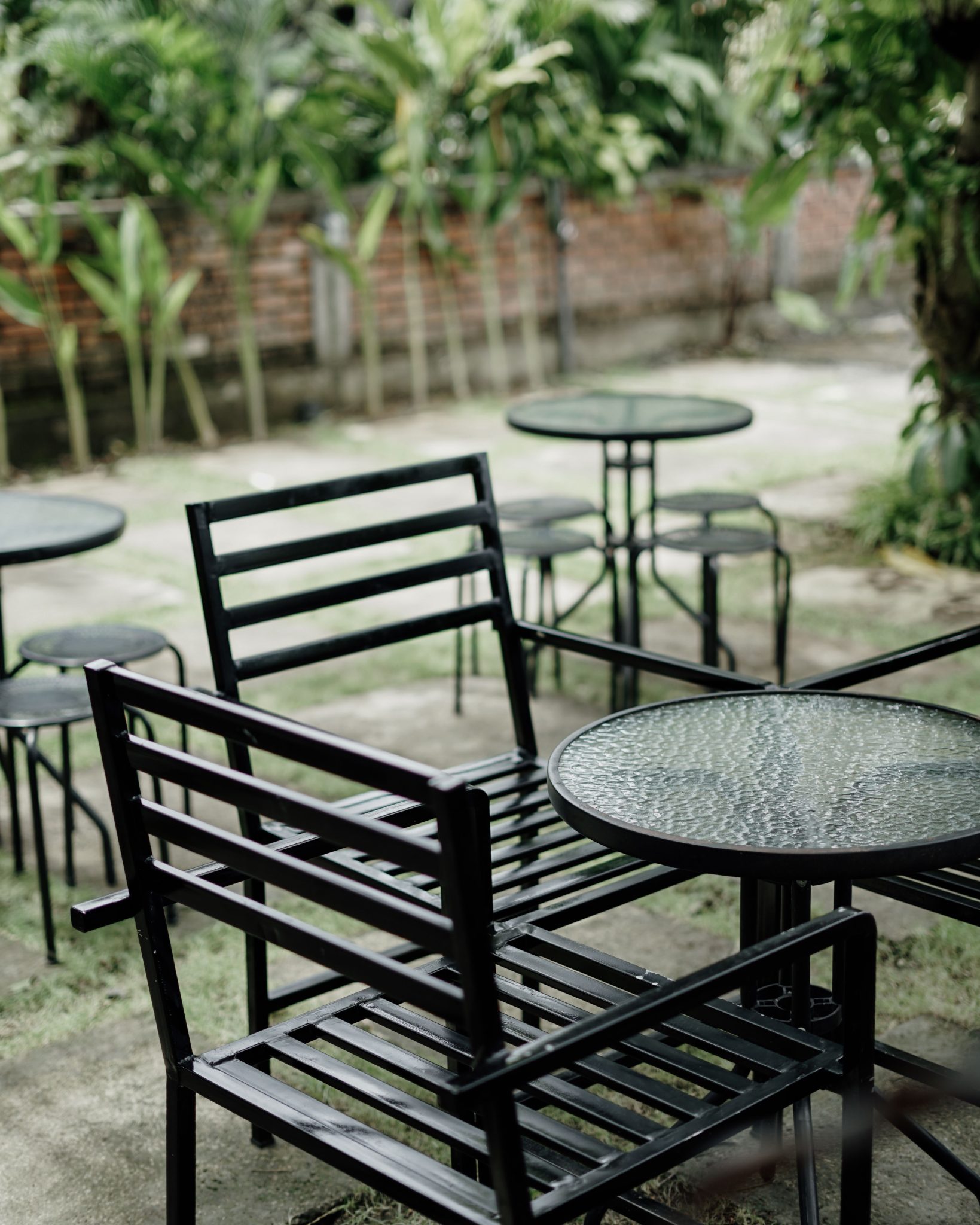 Serene outdoor café setting with empty black metal chairs and glass tables amidst lush green plants.