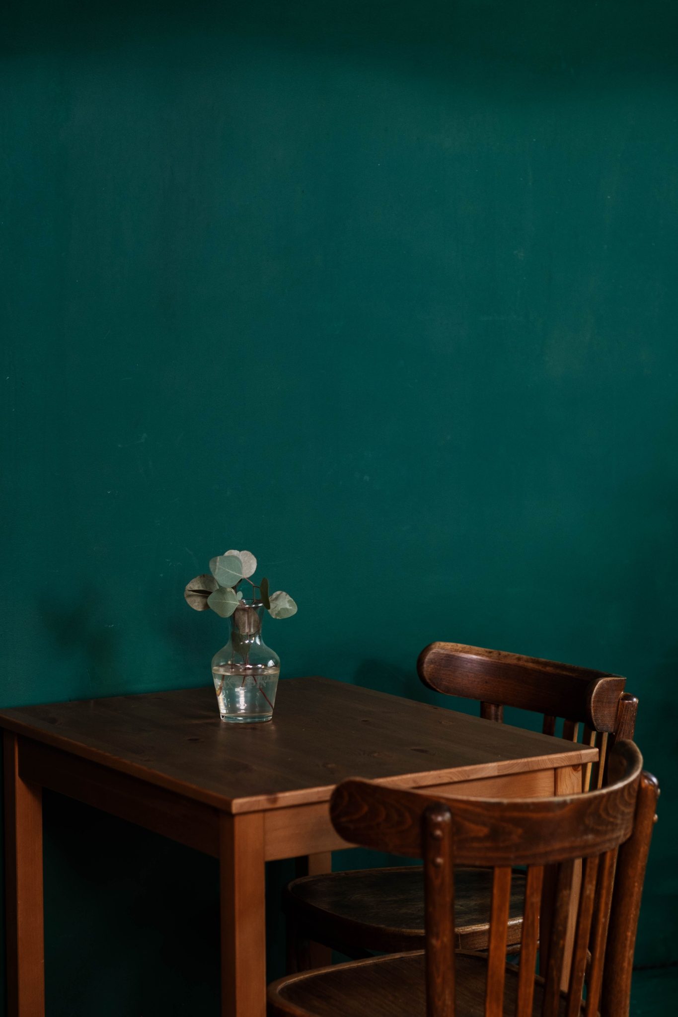 Simple wooden table with a vase and foliage against a teal wall, creating a minimalist aesthetic indoors.