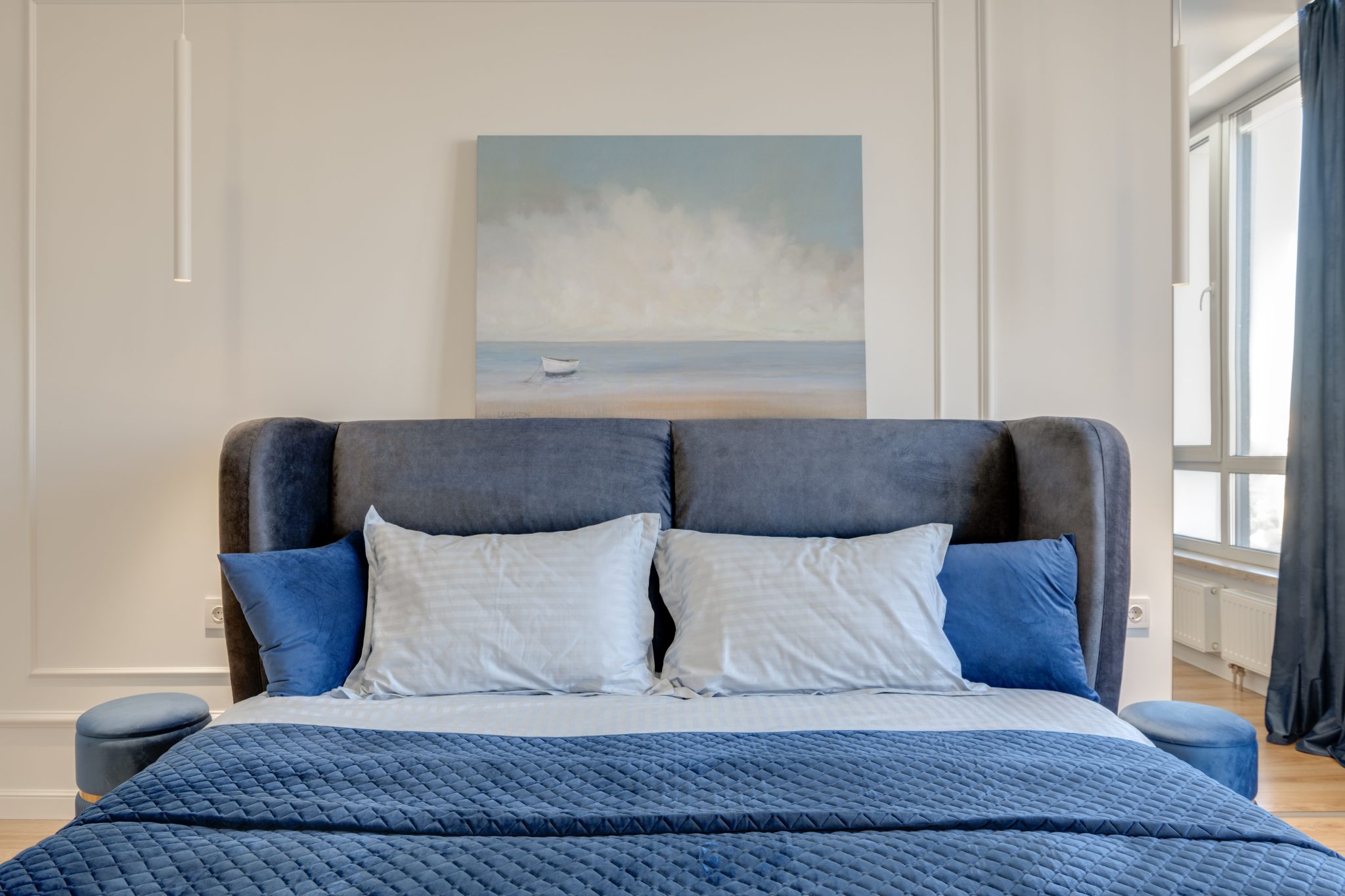 Stylish bedroom interior featuring a blue bedspread, pillows, and an ocean painting. Modern and inviting.