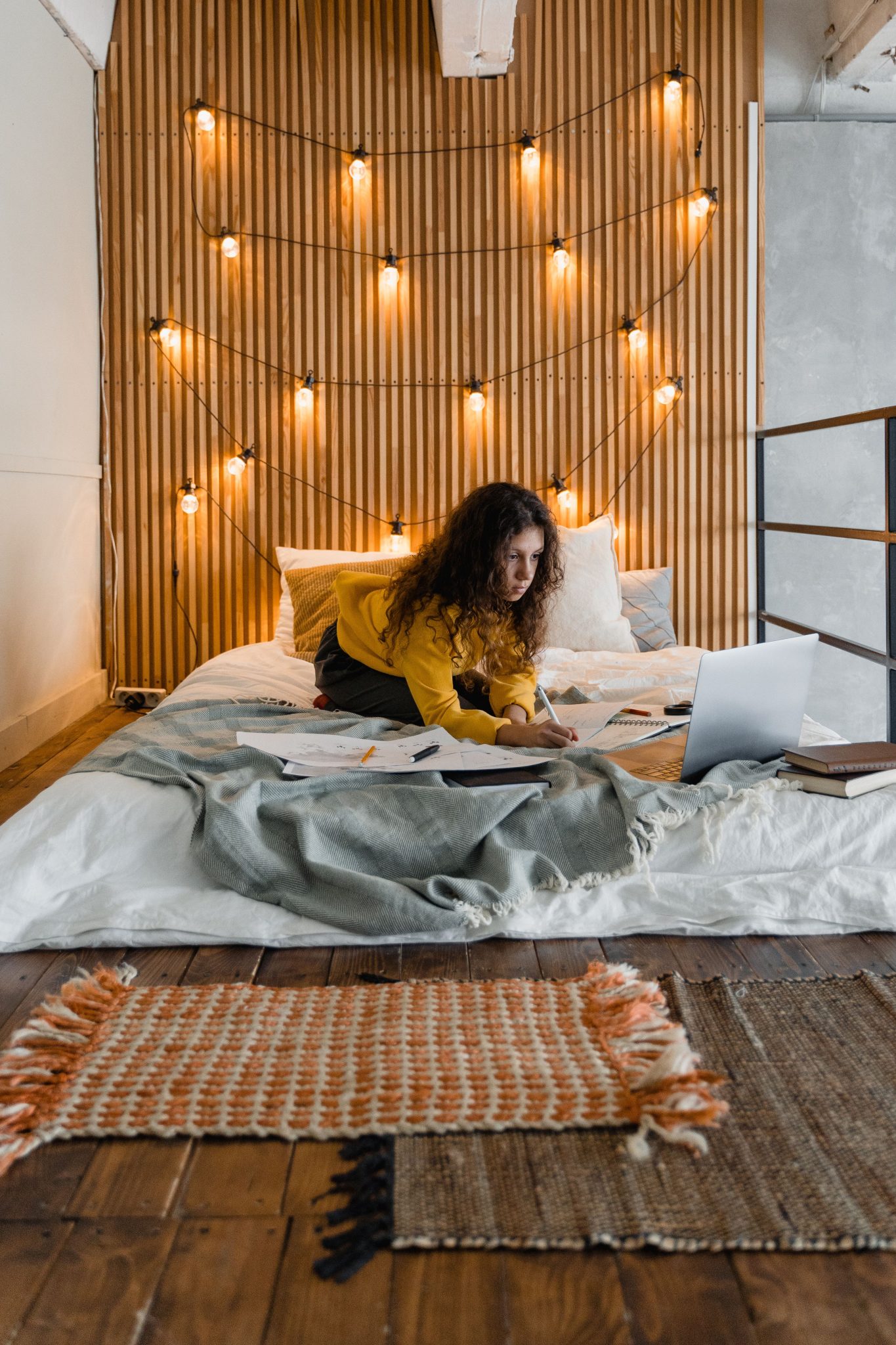 Teenage girl studying on a cozy bed surrounded by books and a laptop in a well-lit room.
