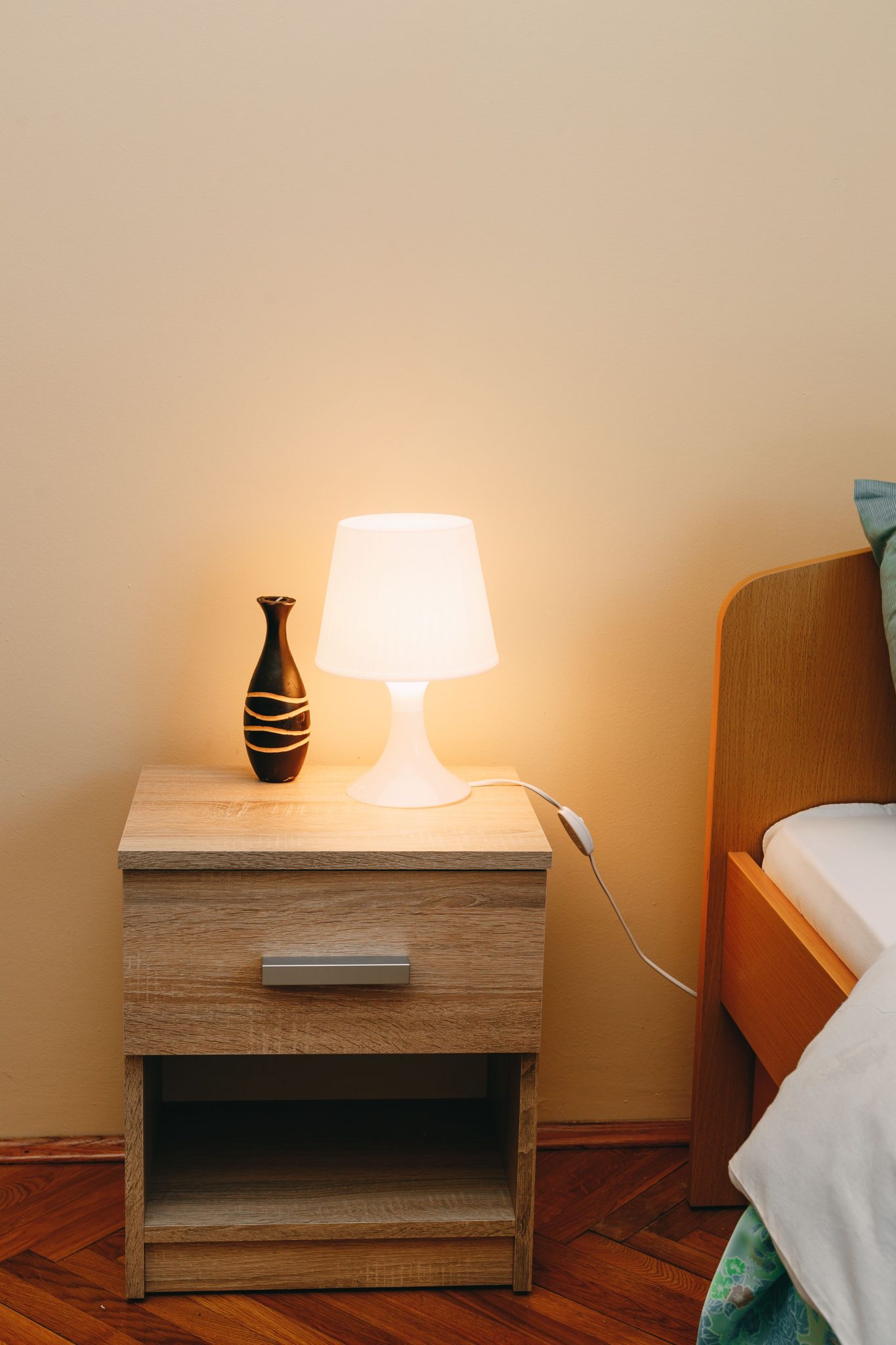 Warm lighting on a wooden nightstand with a lamp and vase next to a comfortable bed. Ideal for home decor inspiration.