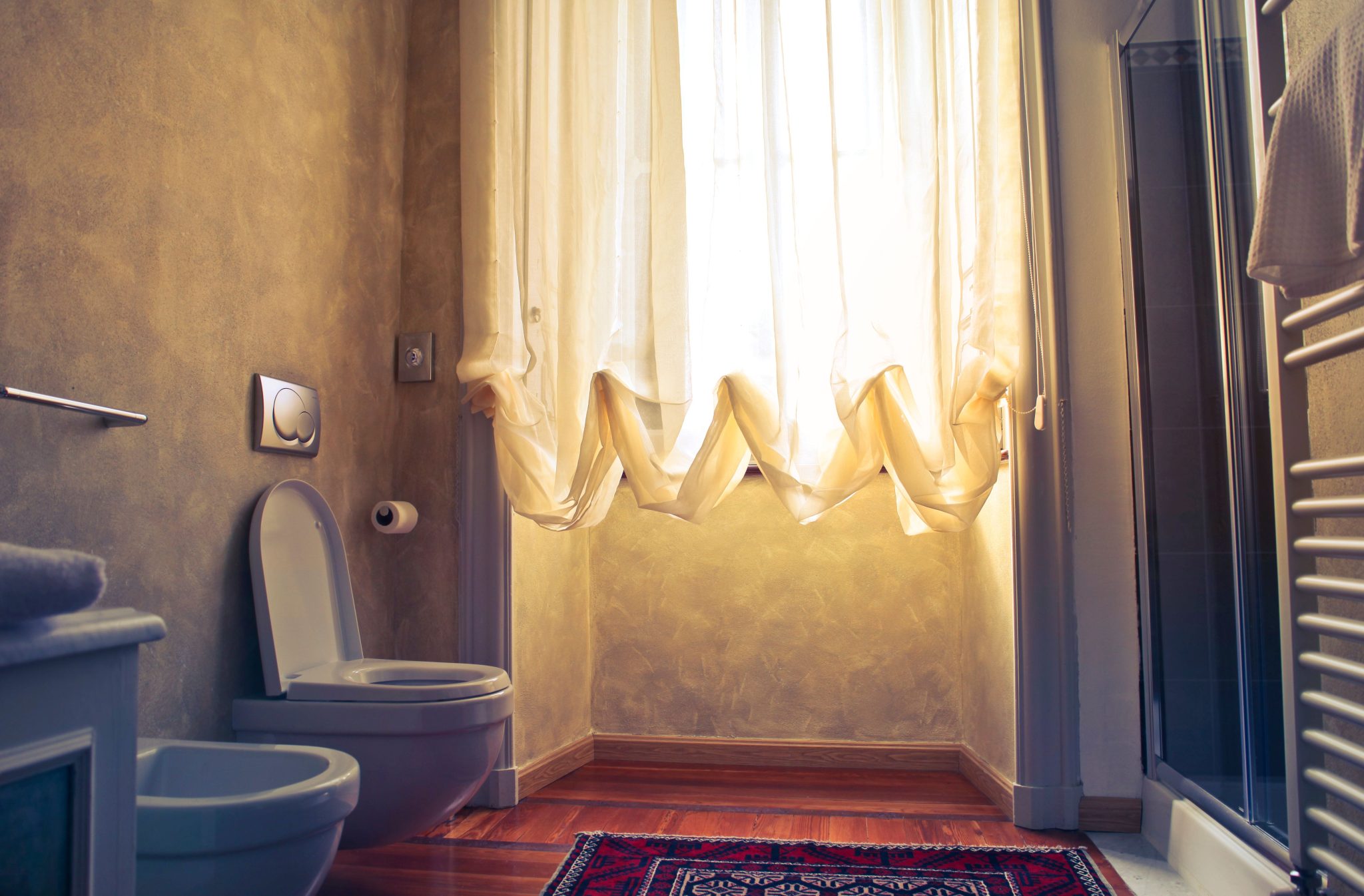 Warmly lit bathroom with a bidet, toilet, and a flowing curtain revealing soft sunlight.