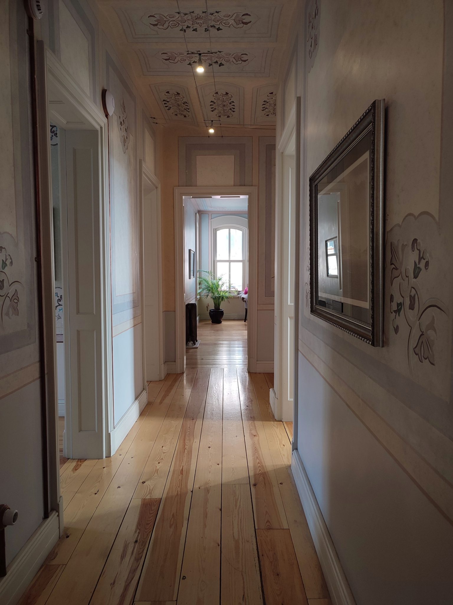 A beautifully lit corridor with wooden floors, ornate walls, and a plant at the end by the window.