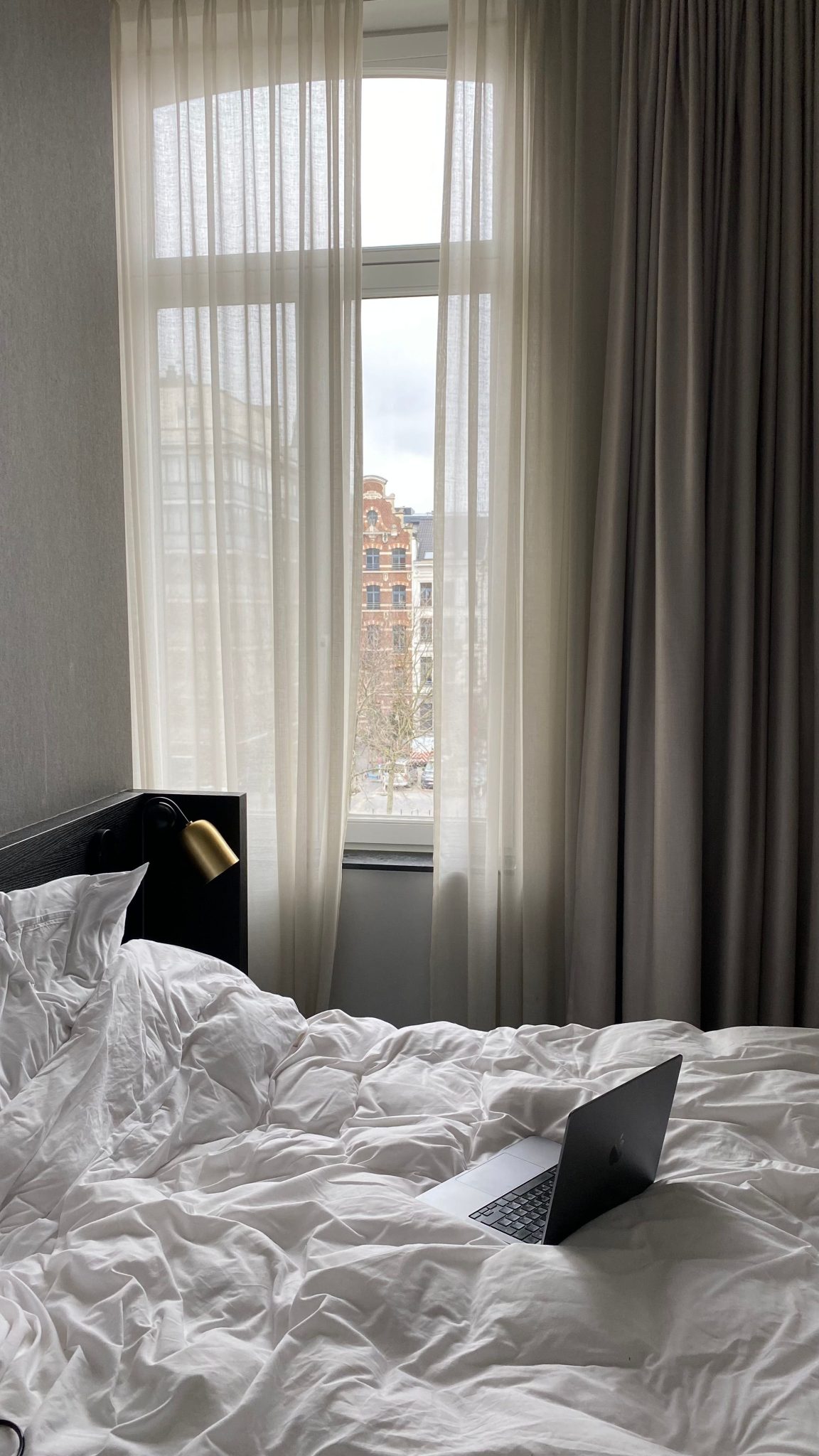 A cozy bedroom setting with a laptop on a bed, featuring a window view of Belgian architecture.