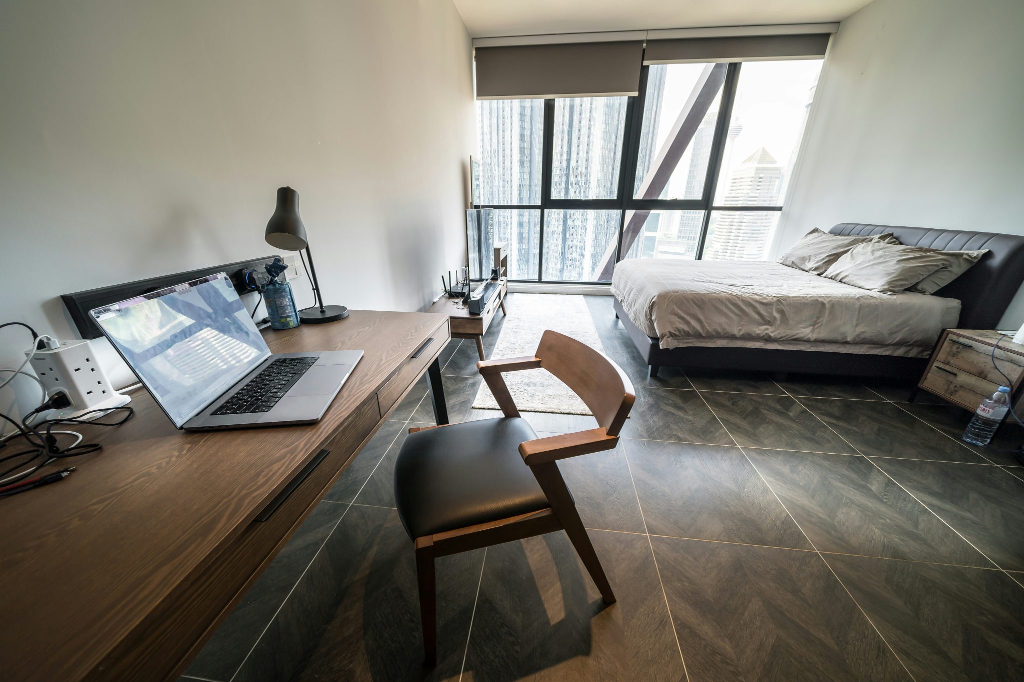 A minimalist studio apartment combining a workspace and bedroom, featuring a wooden desk with a laptop and task lamp alongside a neatly made bed. Large floor-to-ceiling windows frame a sweeping view o