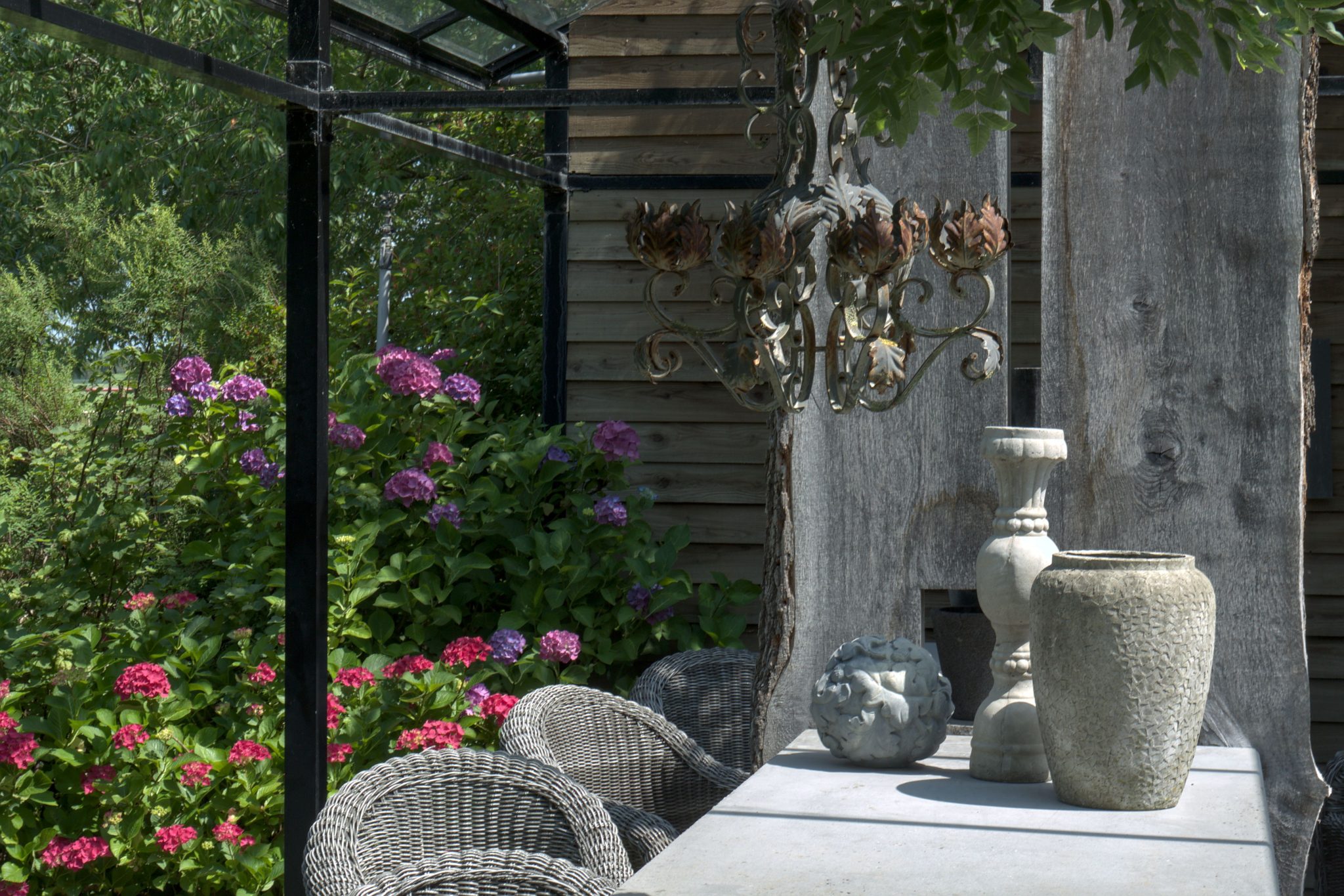 A rustic outdoor garden table adorned with clay pottery and a chandelier amidst vibrant hydrangeas.