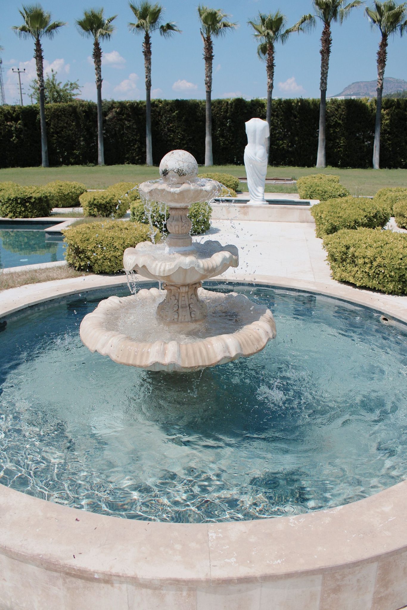 A serene garden fountain surrounded by lush greenery and statues, under a clear blue sky.