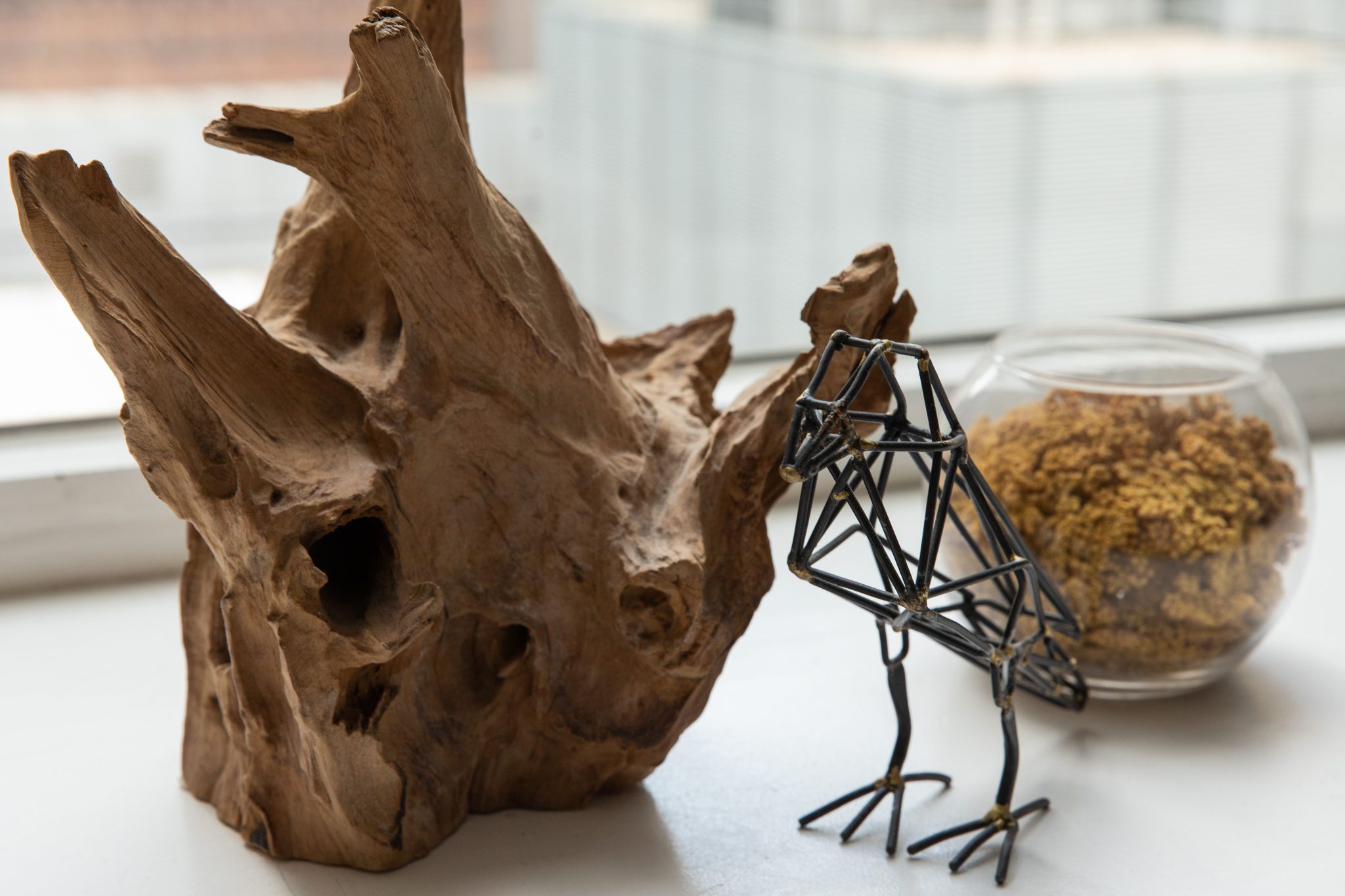 A stunning wire bird sculpture beside natural driftwood and lichen in a glass bowl on a windowsill.