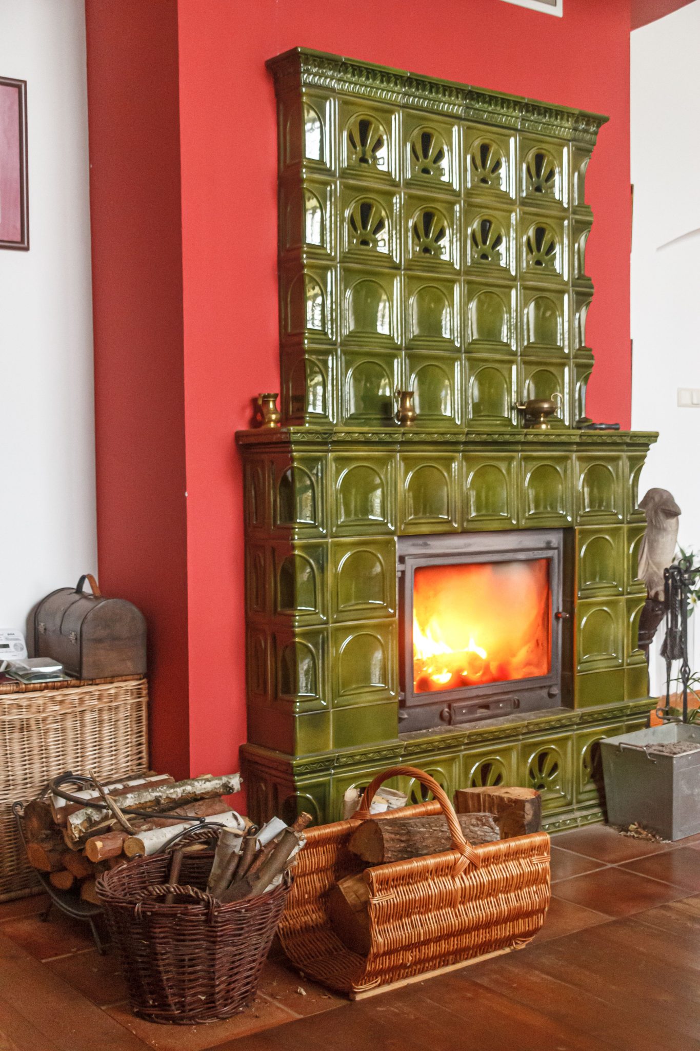 A vintage green tiled fireplace with a warm fire, surrounded by wicker baskets and wooden floor for a cozy setting.