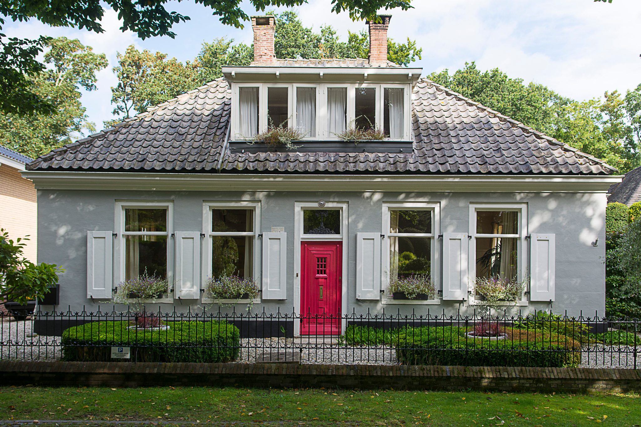Beautiful cottage with white shutters and bright red door, surrounded by lush greenery.